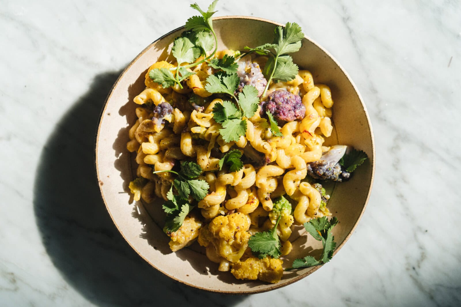 A bowl of cavatappi pasta with cauliflower, purple vegetables, and fresh cilantro on a marble surface. - Hetty Lui McKinnon
