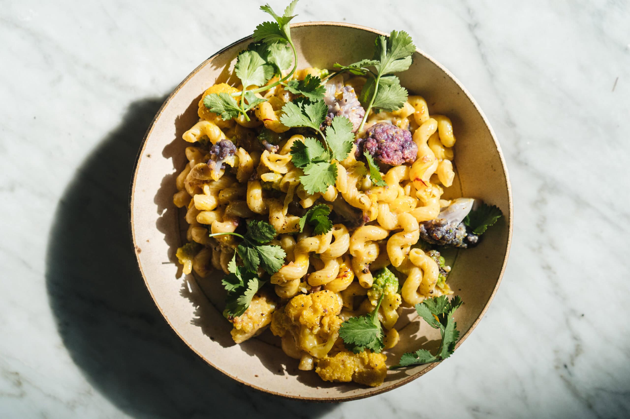 A bowl of cavatappi pasta with cauliflower, purple vegetables, and fresh cilantro on a marble surface. - Hetty Lui McKinnon