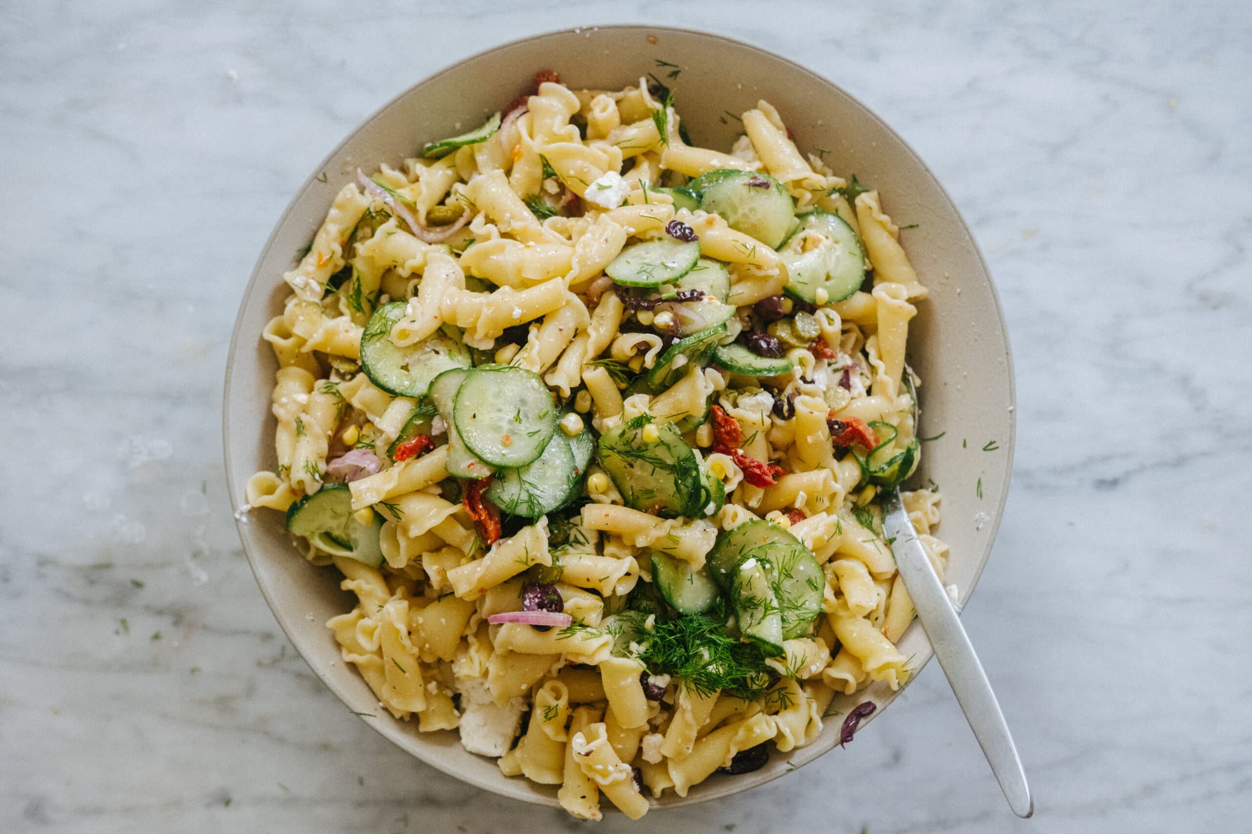 A bowl of pasta salad with sliced cucumbers, olives, red onion, feta cheese, dill, and a fork, on a marble surface.