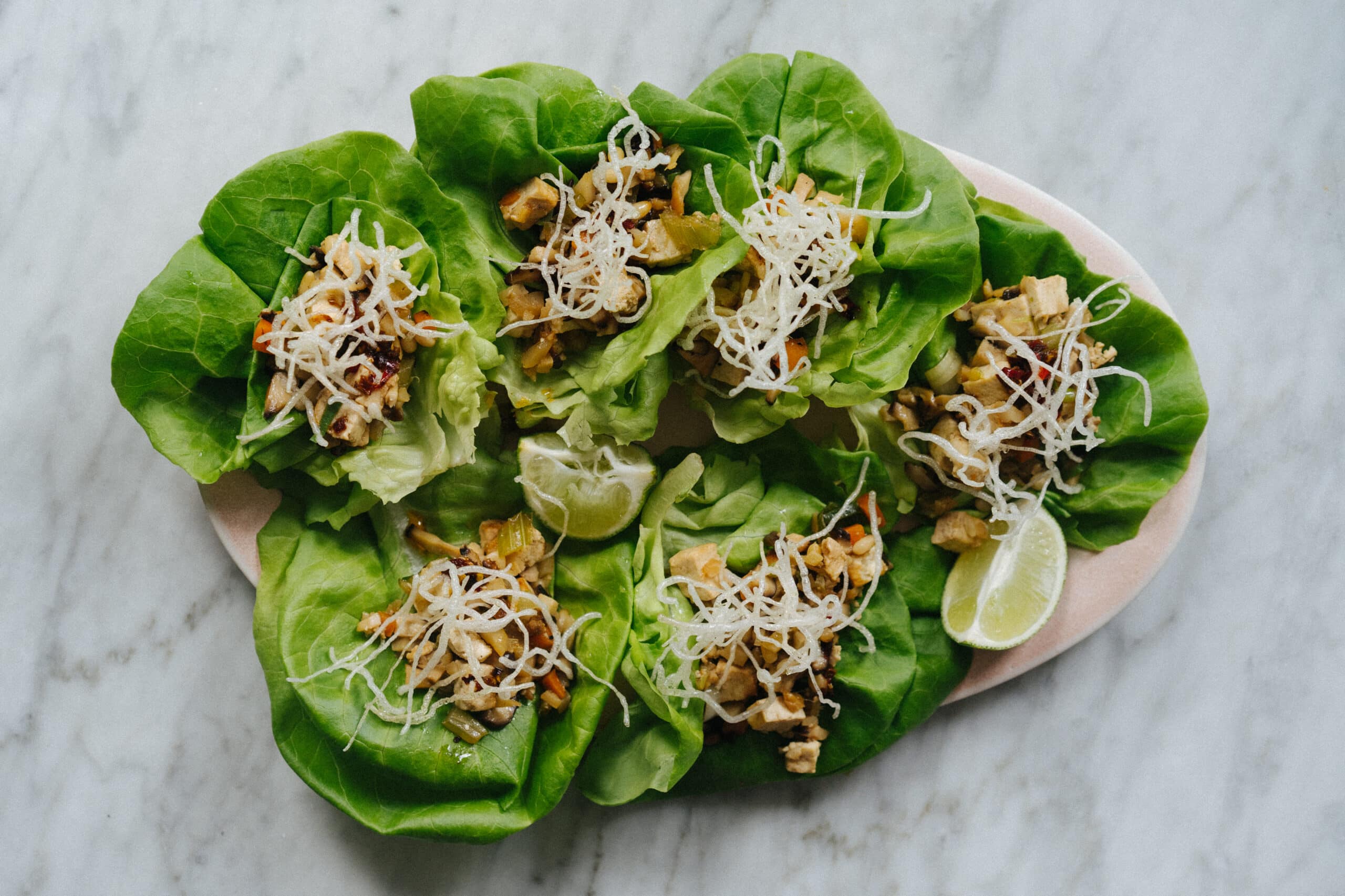 A plate of lettuce wraps filled with a diced vegetable and protein mixture, topped with shredded white cheese and served with lime wedges on a marble surface.