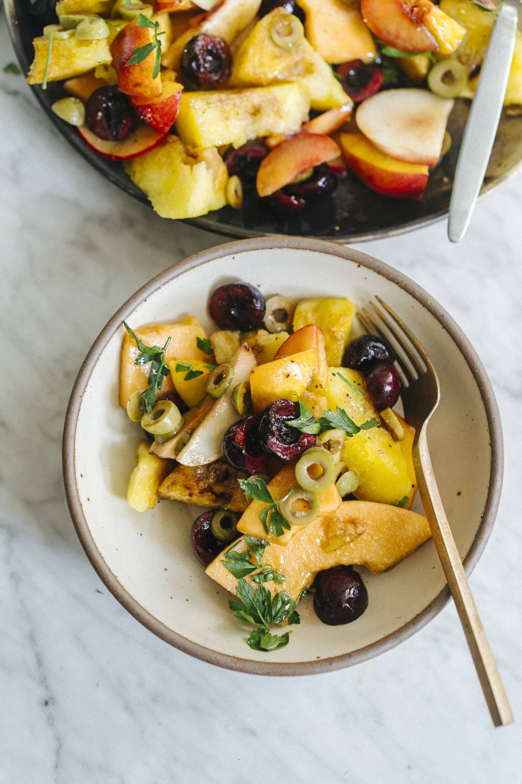 A bowl of mixed fruit salad with cantaloupe, cherries, green olives, fresh herbs, and a gold fork on a marble surface. Another bowl of the salad is partially visible above.