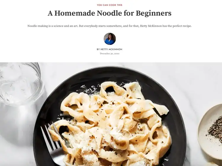 A plate of homemade fettuccine noodles topped with grated cheese, served with a fork on a black plate, next to a small dish of pepper.