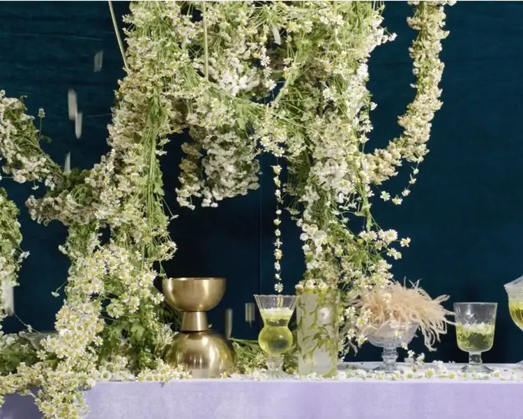 A table with green drinks in glassware, a gold chalice, and vases is decorated with cascading white and yellow flowers on a dark blue background.