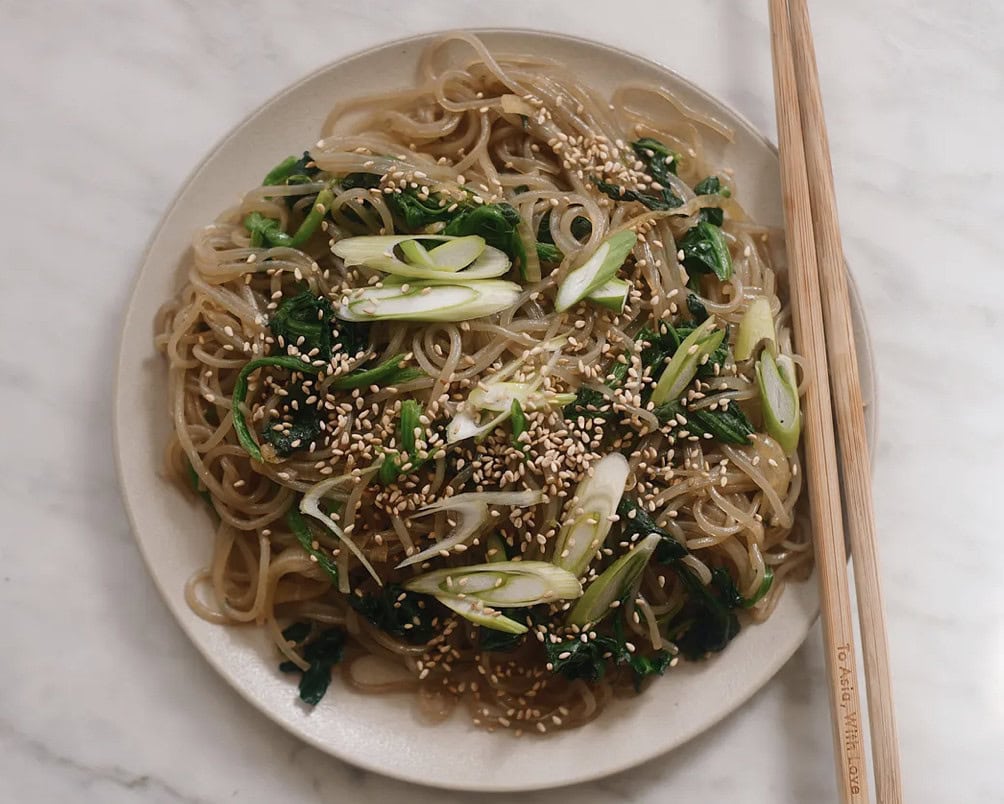 A plate of glass noodles with sautéed greens, topped with sliced green onions and sesame seeds, next to a pair of wooden chopsticks.