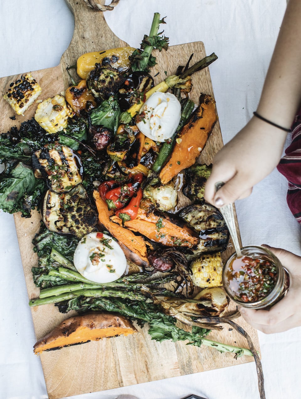 A wooden board with grilled vegetables, including corn, asparagus, peppers, and sweet potato, topped with mozzarella balls. A hand spoons dressing from a jar onto the food. - Hetty Lui McKinnon