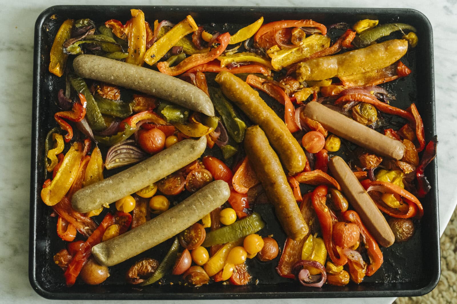 Sheet pan with cooked sausages, sliced bell peppers, cherry tomatoes, red onion, and olives arranged on a black baking tray. - Hetty Lui McKinnon