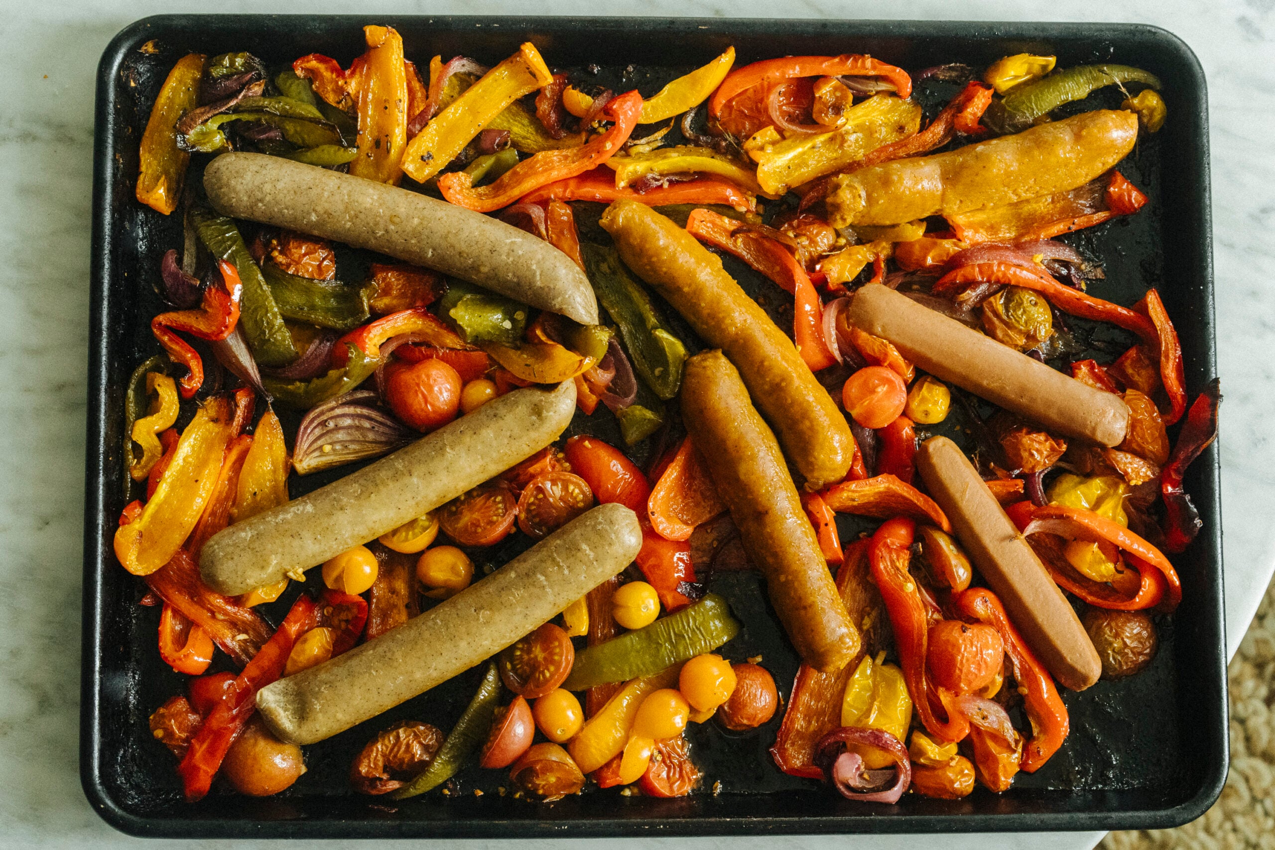 Sheet pan with cooked sausages, sliced bell peppers, cherry tomatoes, red onion, and olives arranged on a black baking tray. - Hetty Lui McKinnon