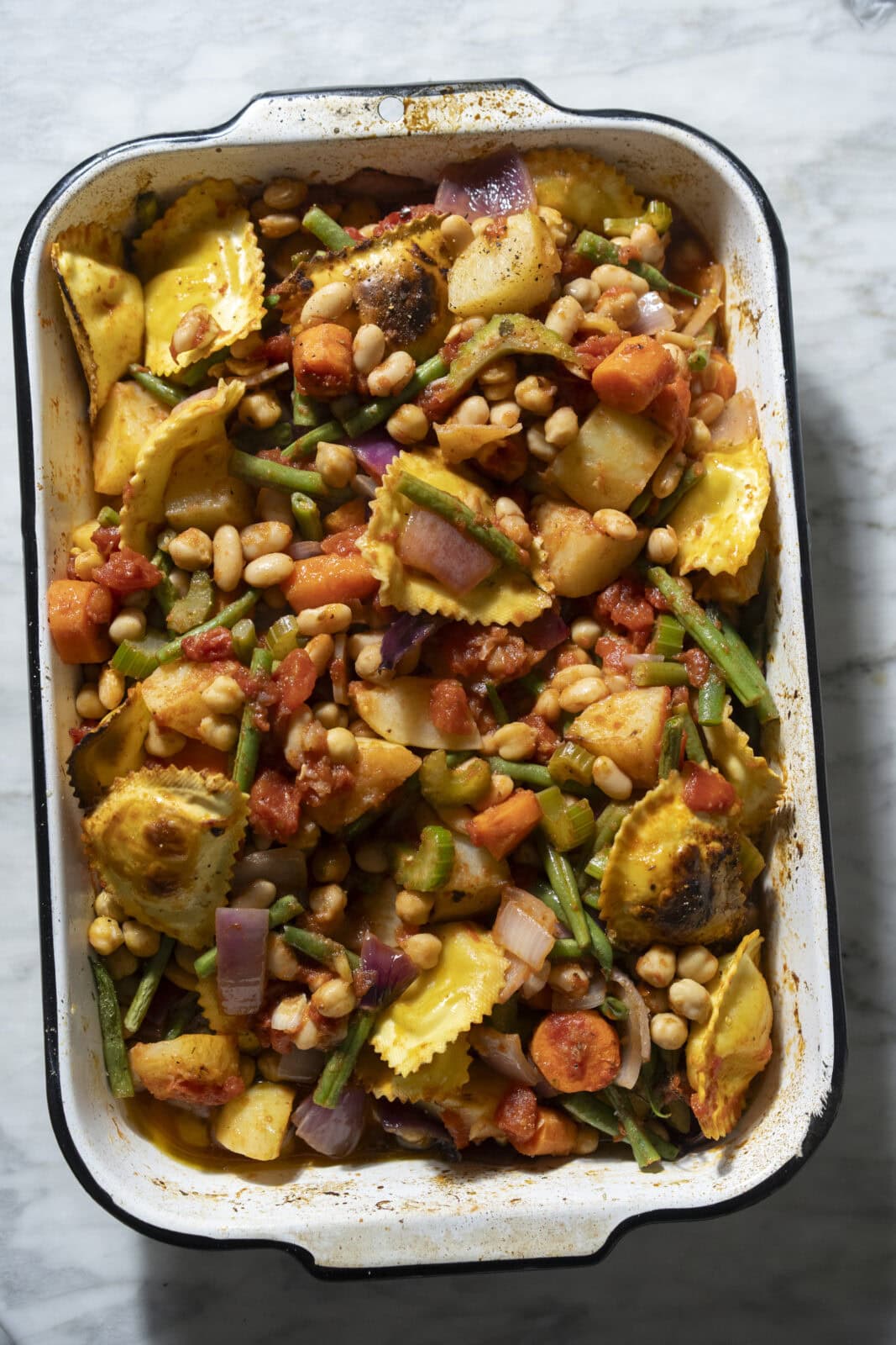 A baking dish filled with roasted ravioli, chickpeas, carrots, potatoes, green beans, tomatoes, and red onions on a marble surface.