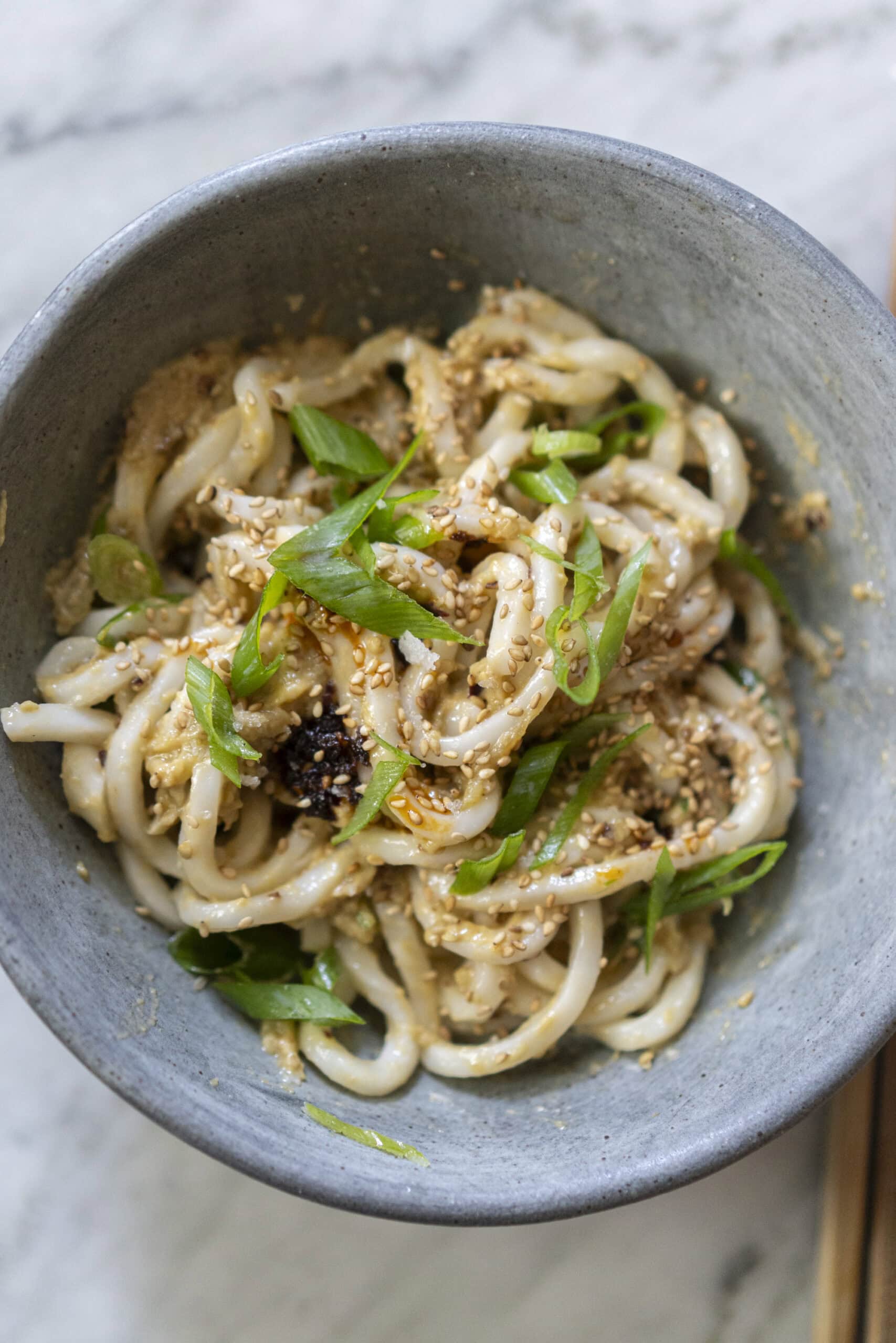 A bowl of udon noodles topped with sliced green onions, sesame seeds, and sauce, served in a gray bowl on a marble surface.