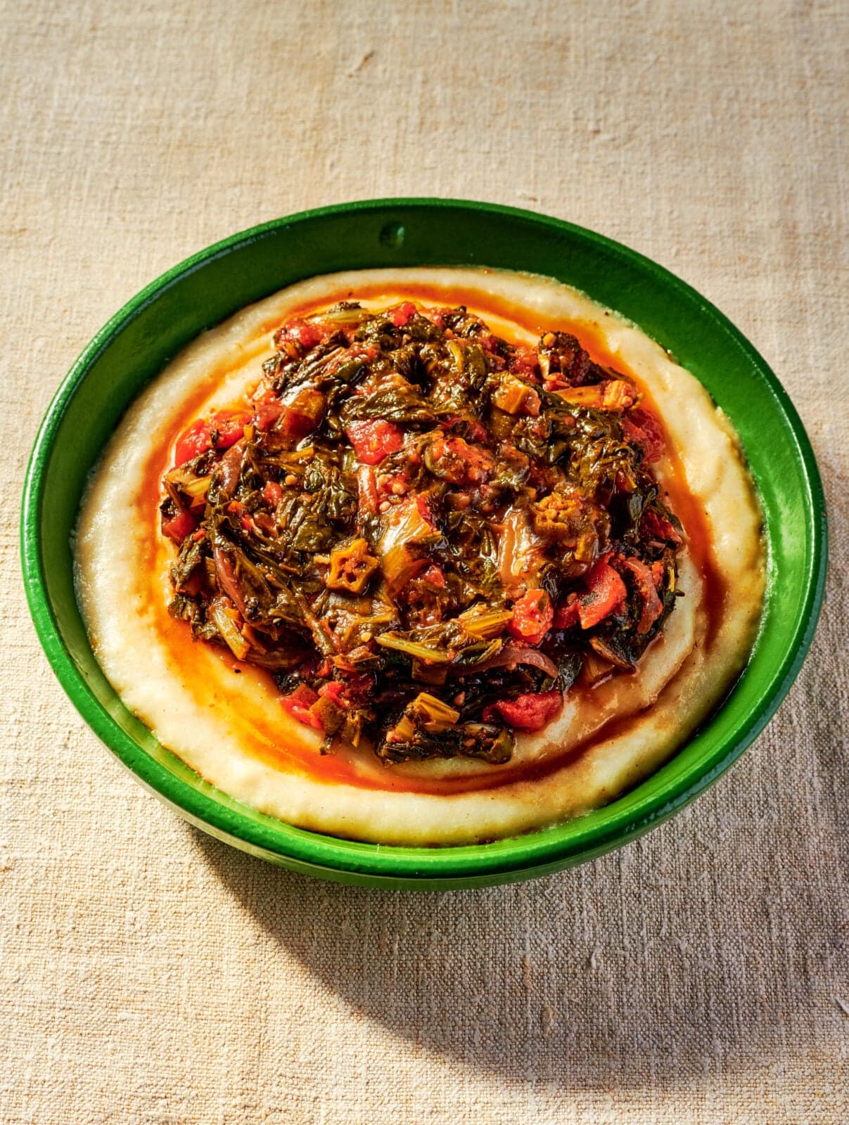 A green bowl filled with creamy polenta topped with a cooked mixture of leafy greens, tomatoes, and vegetables on a beige surface.