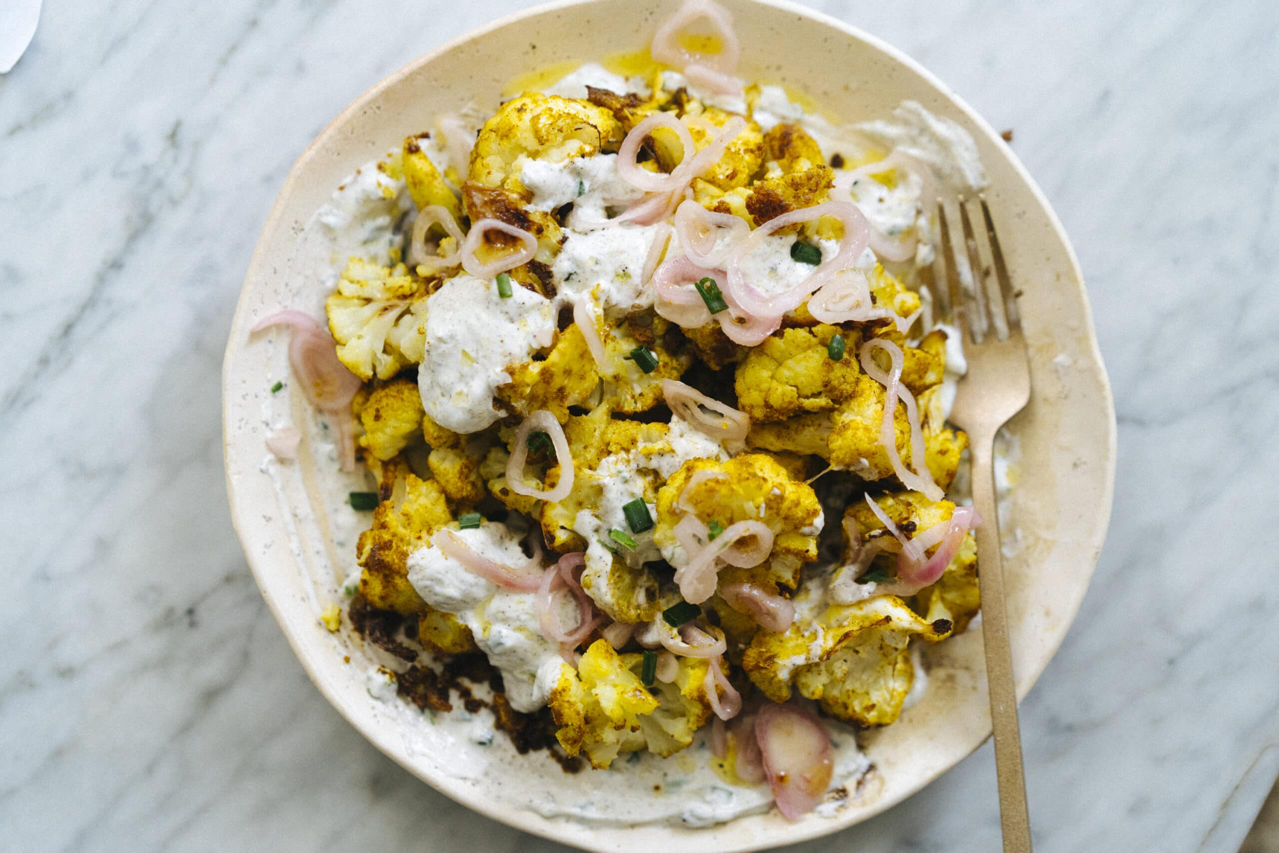 A plate of roasted cauliflower topped with white sauce, pickled onions, and chopped herbs, with a fork on the side.
