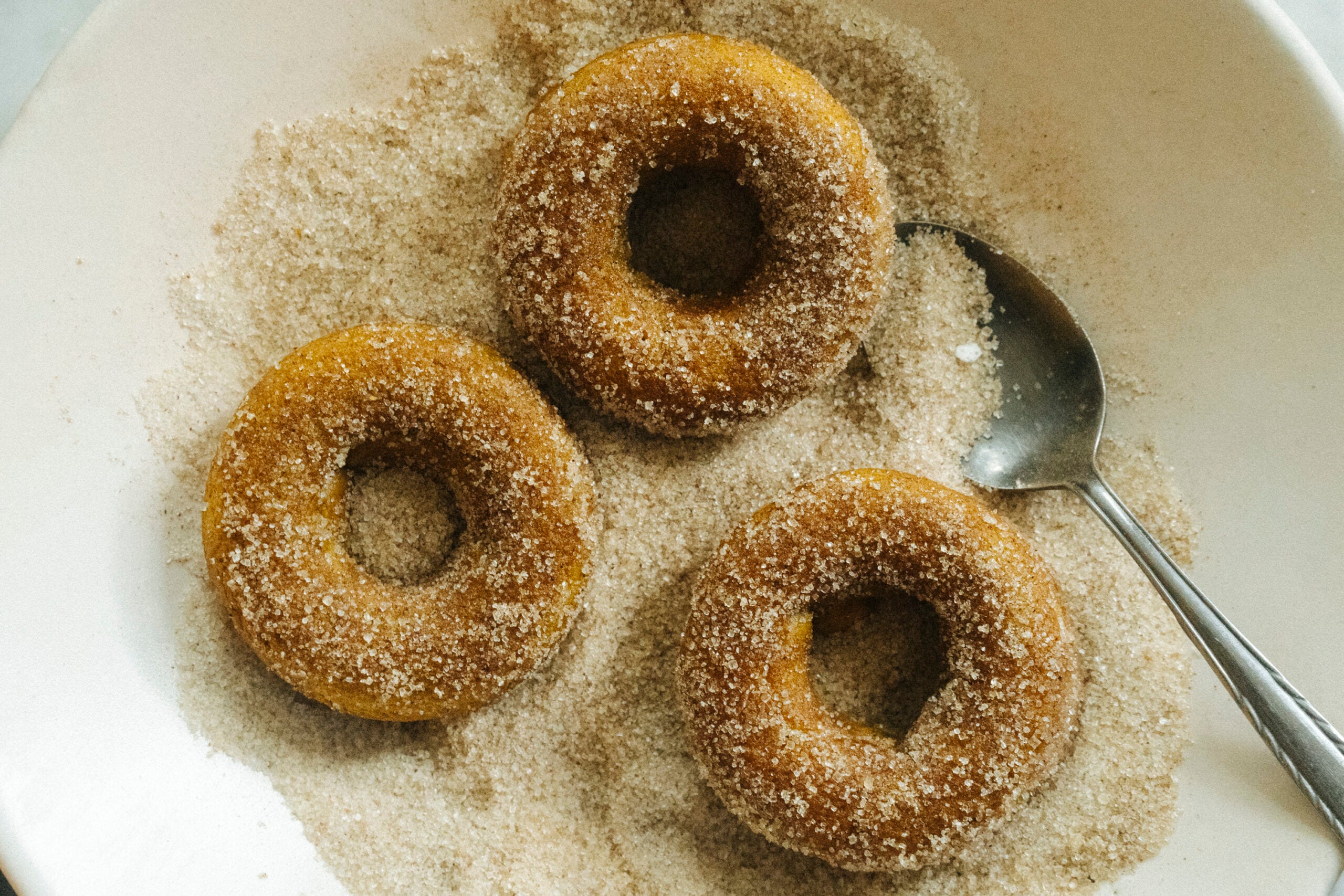 Three sugar-coated donuts in a bowl of sugar with a metal spoon. - Hetty Lui McKinnon
