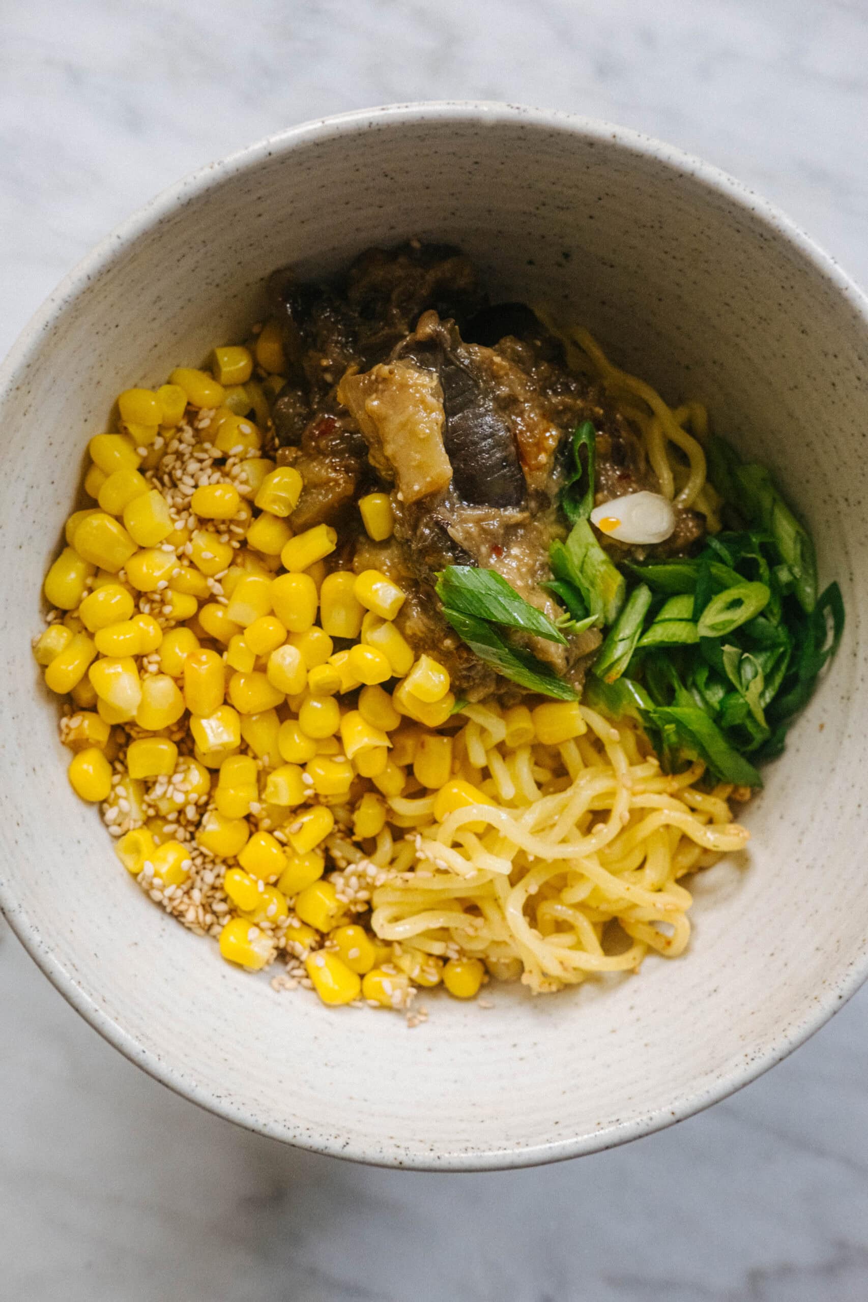 A bowl of noodles topped with corn, sesame seeds, green onions, and a portion of stewed meat or vegetables.