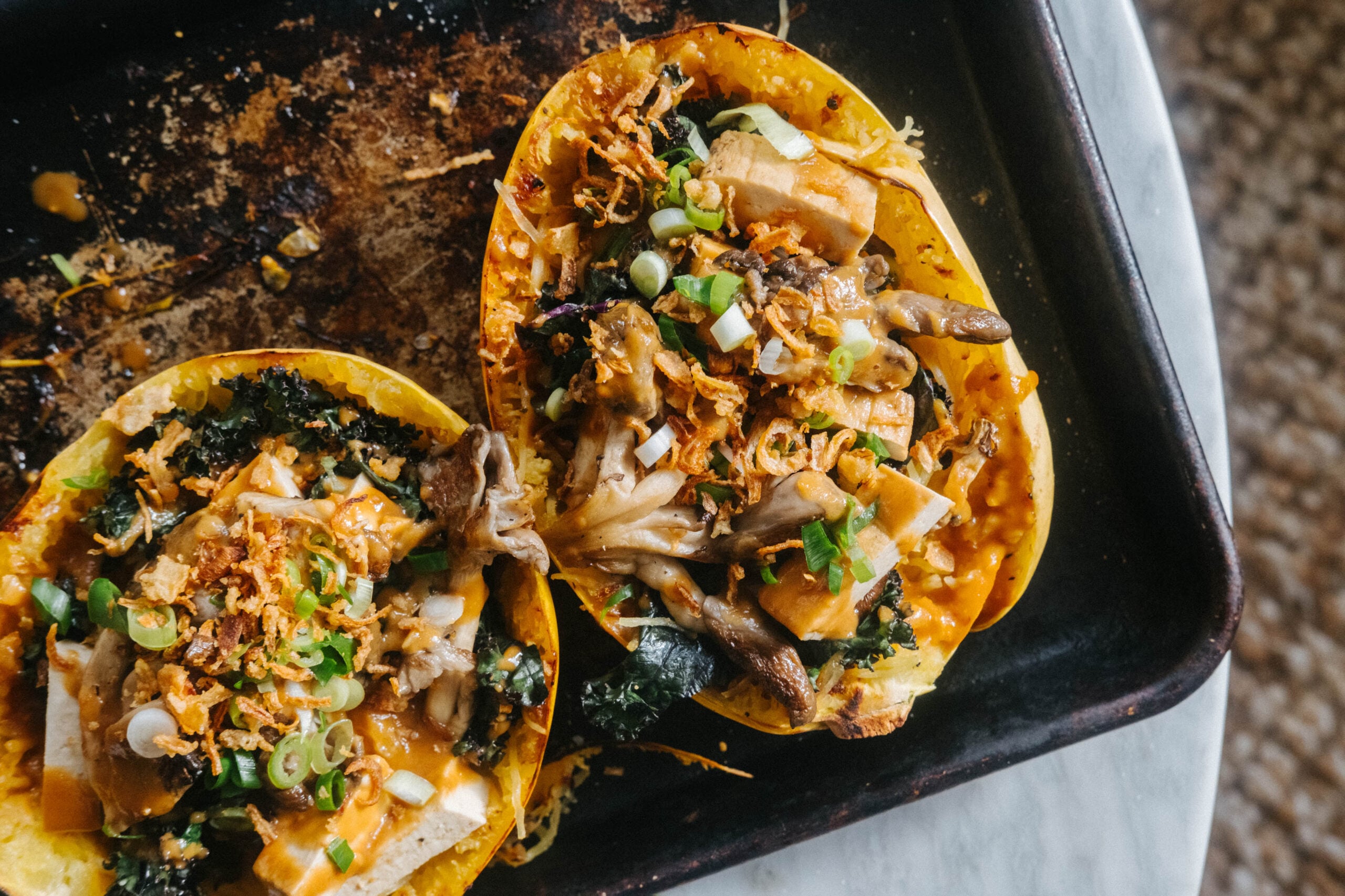 Two roasted spaghetti squash halves topped with tofu, mushrooms, kale, crispy onions, and green onions on a baking sheet. - Hetty Lui McKinnon