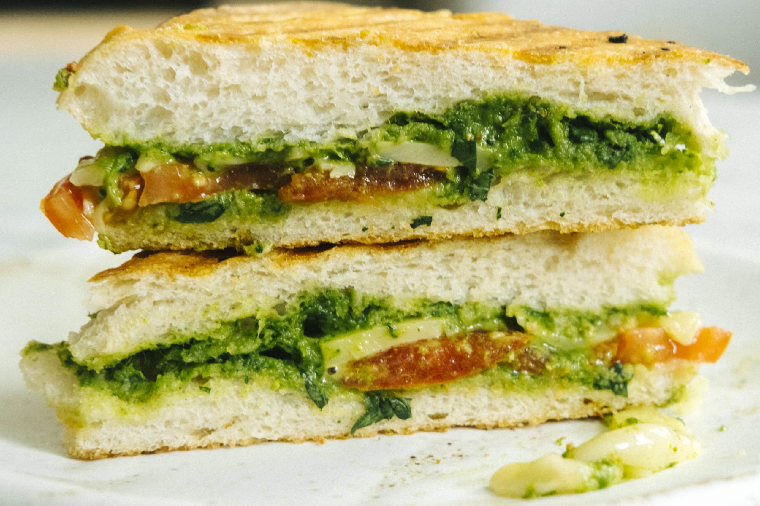 A close-up of a sandwich cut in half, filled with green pesto, slices of tomato, cheese, and leafy greens on white bread.