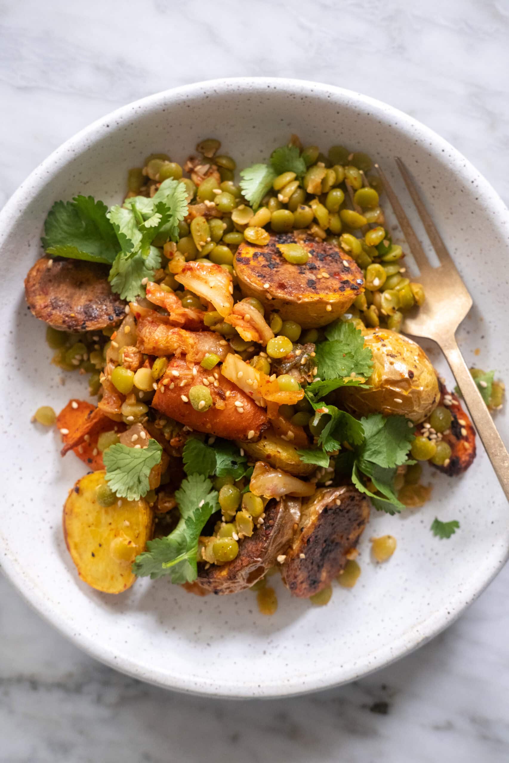 A white bowl with roasted potatoes, green lentils, kimchi, fresh cilantro, and a gold fork on a marble surface.