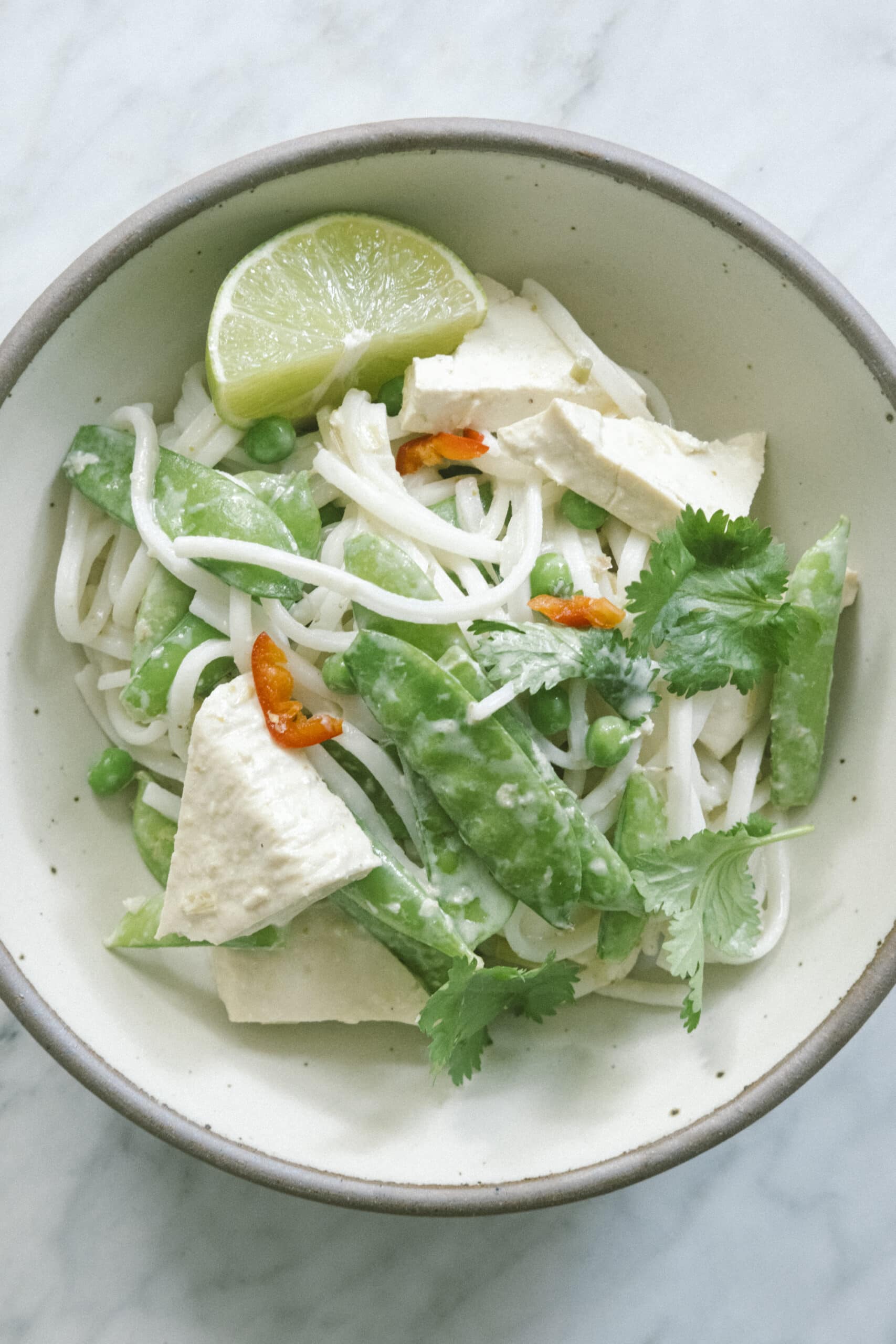 A bowl of rice noodles with tofu, snap peas, peas, cilantro, red chili slices, and a lime wedge on a white surface.