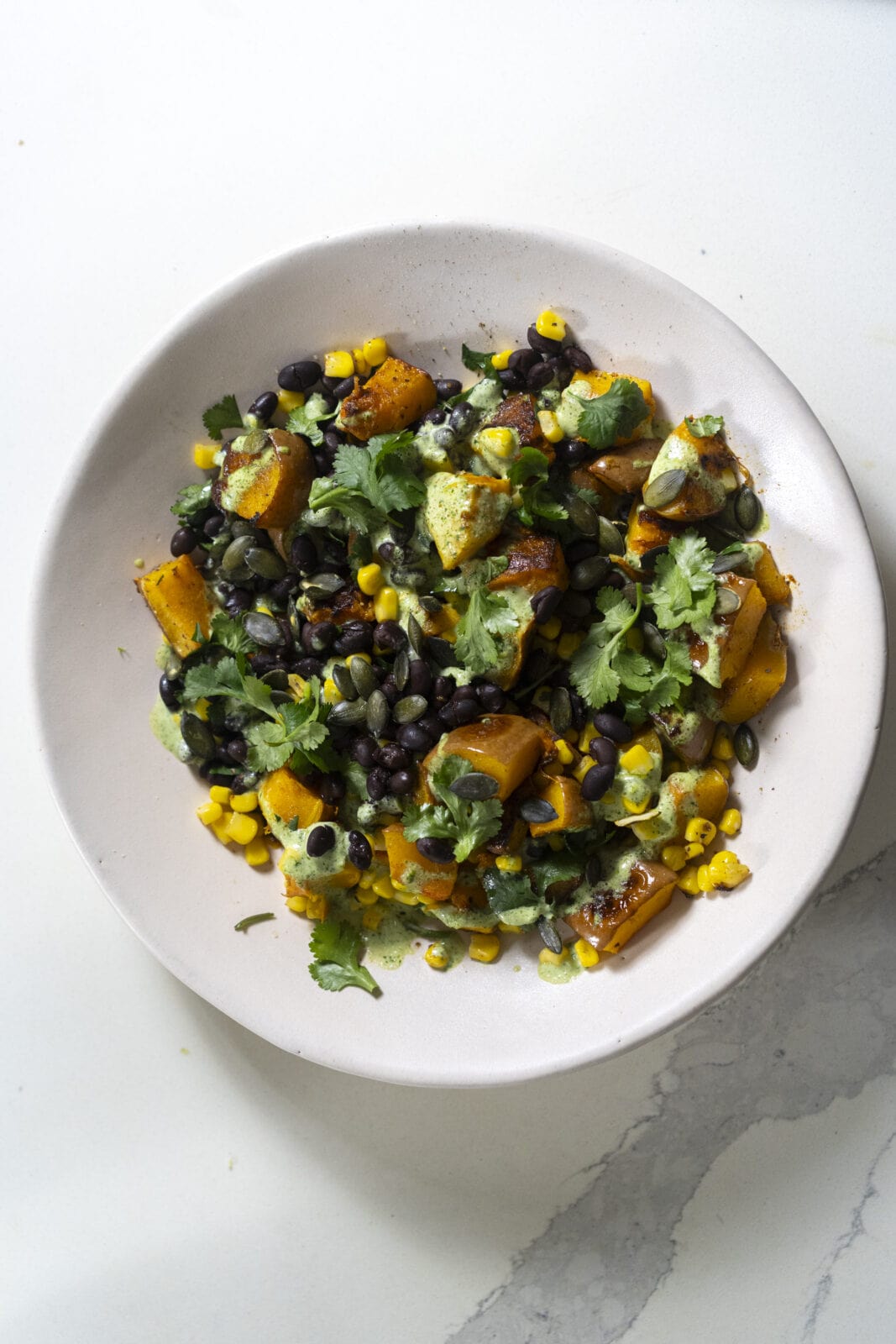 A white bowl filled with a salad of roasted sweet potatoes, black beans, corn, chopped cilantro, and green dressing on a white surface.