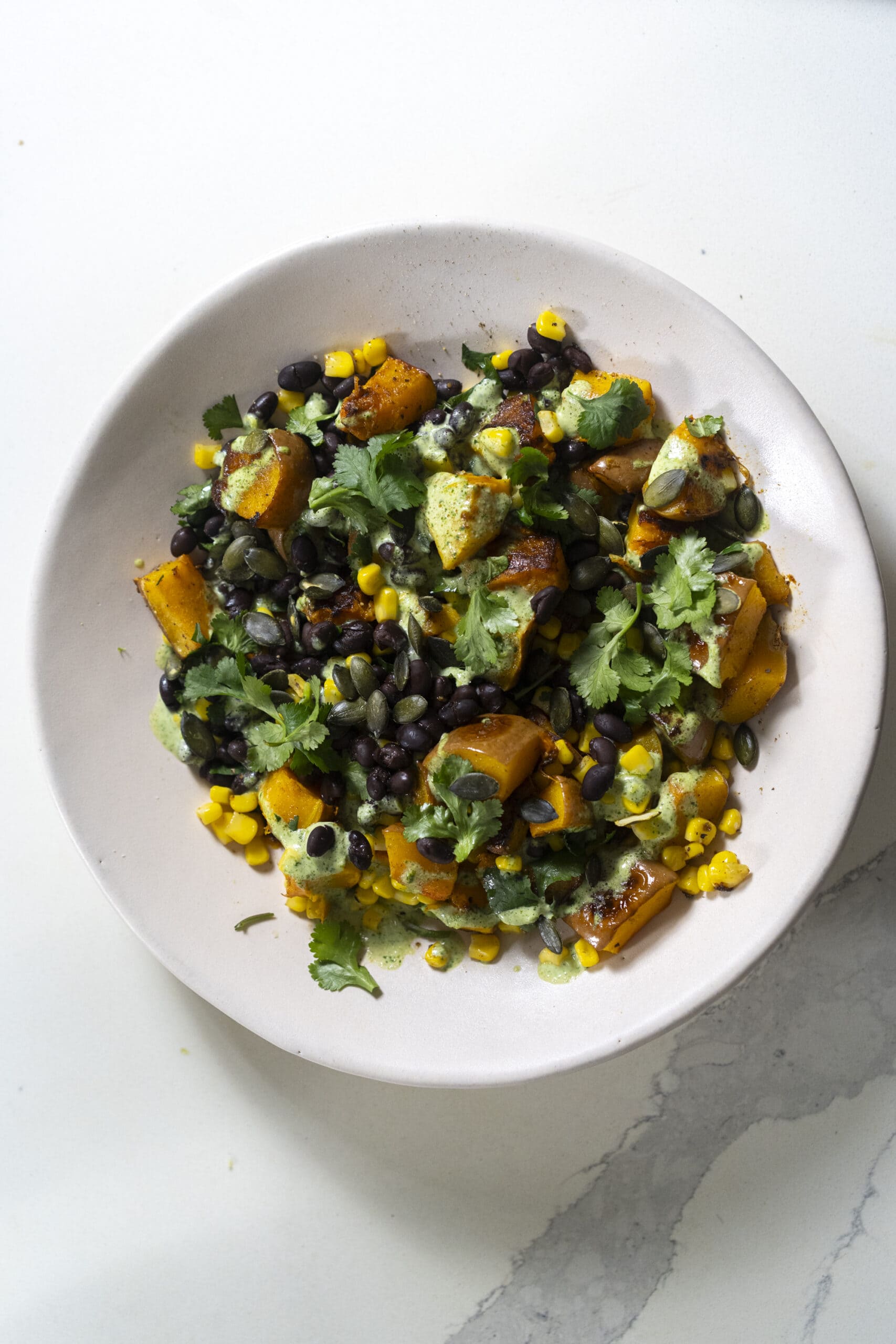 A white bowl filled with a salad of roasted sweet potatoes, black beans, corn, chopped cilantro, and green dressing on a white surface.