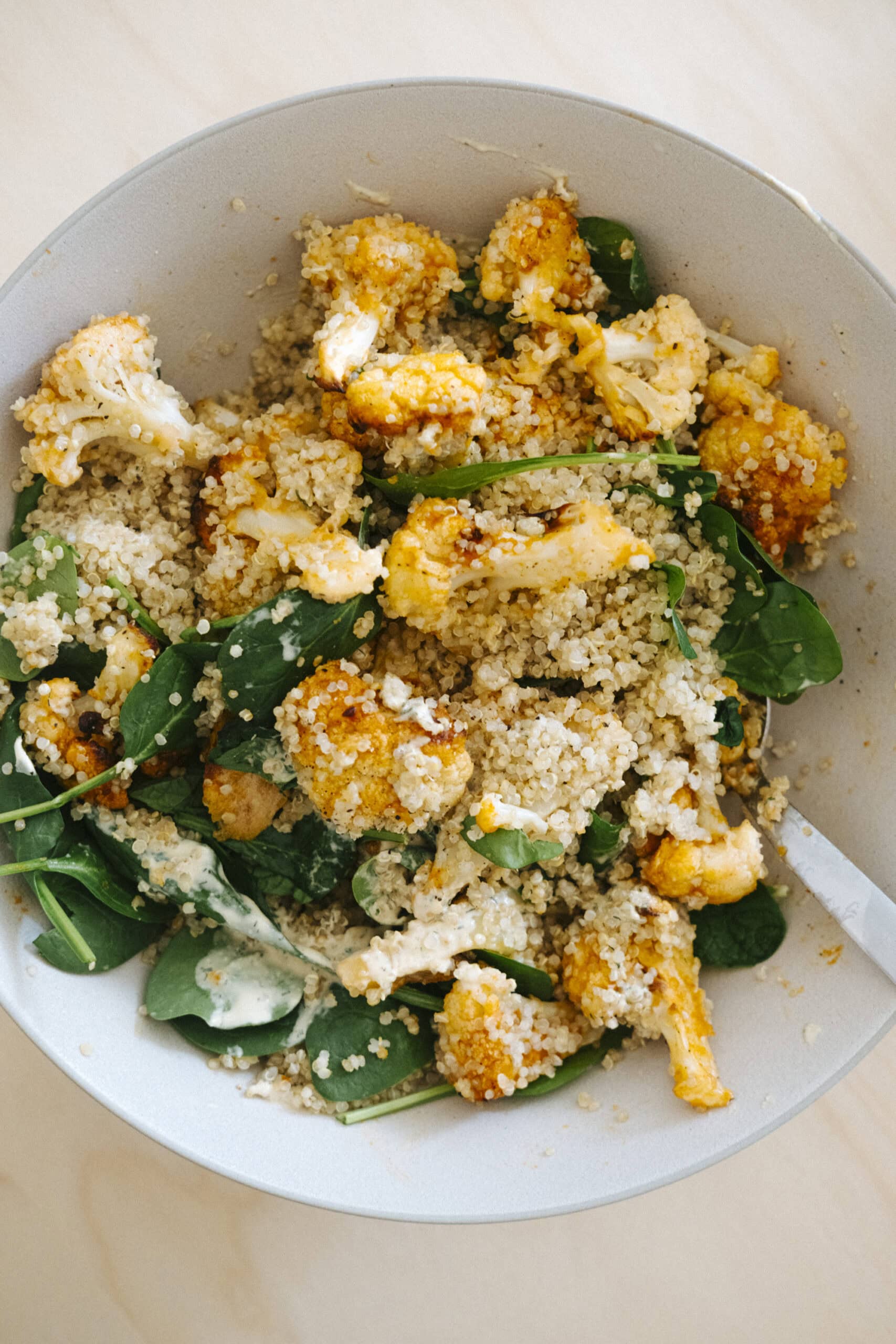 A bowl of quinoa salad with roasted cauliflower, fresh spinach, and a light creamy dressing, served with a fork.