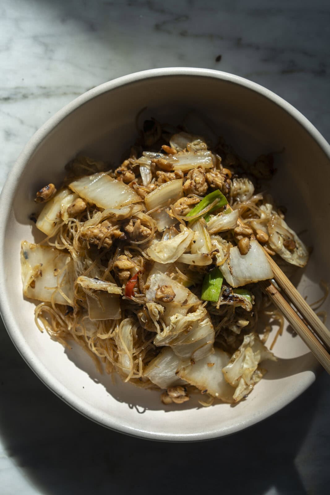 A bowl of stir-fried rice noodles with chopped cabbage, green onions, and pieces of meat, with a pair of chopsticks resting on the side.