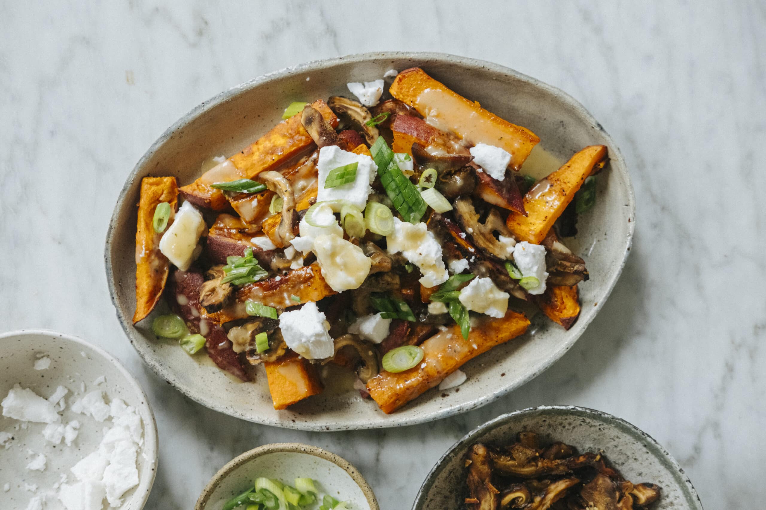 Oval plate with roasted sweet potato wedges, topped with crumbled cheese, sautéed mushrooms, chopped green onions, and herbs on a marble surface.
