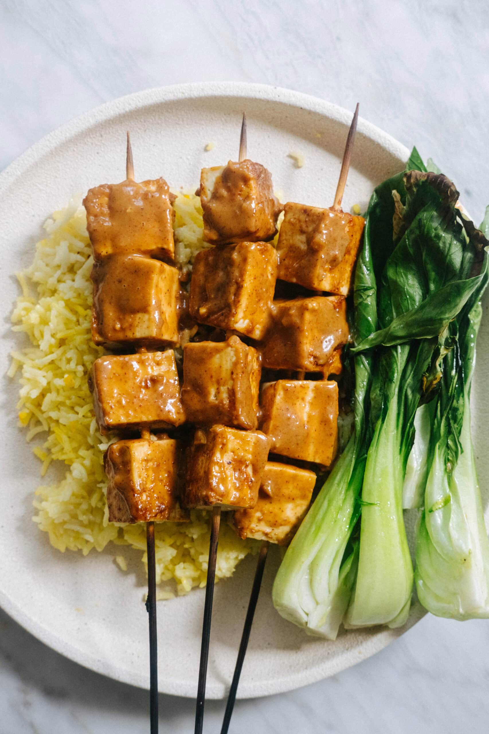 Three skewers of marinated tofu served on yellow rice with steamed bok choy on a white plate.