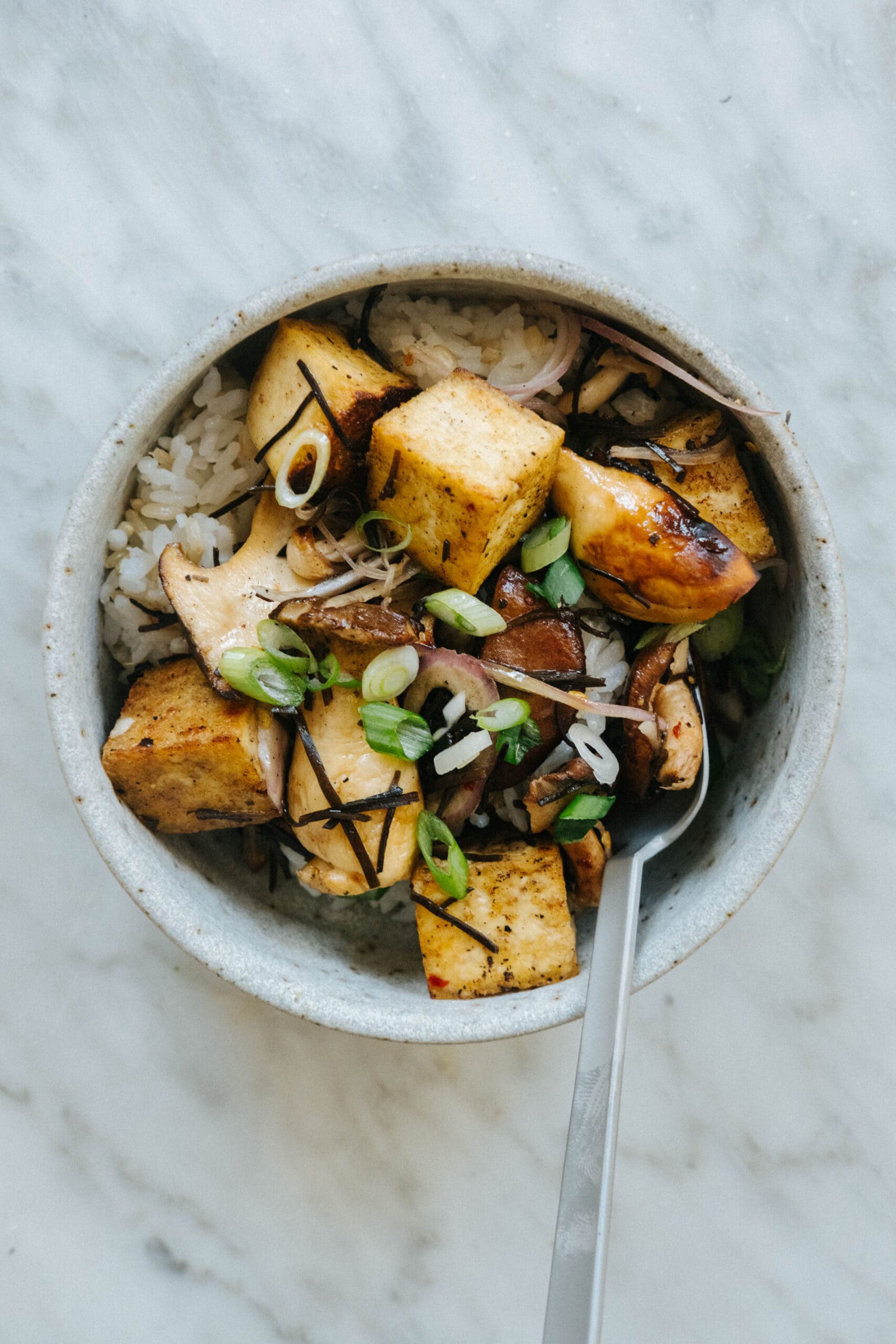 A bowl of rice topped with tofu cubes, sliced mushrooms, green onions, and thin seaweed strips, with a spoon resting inside.