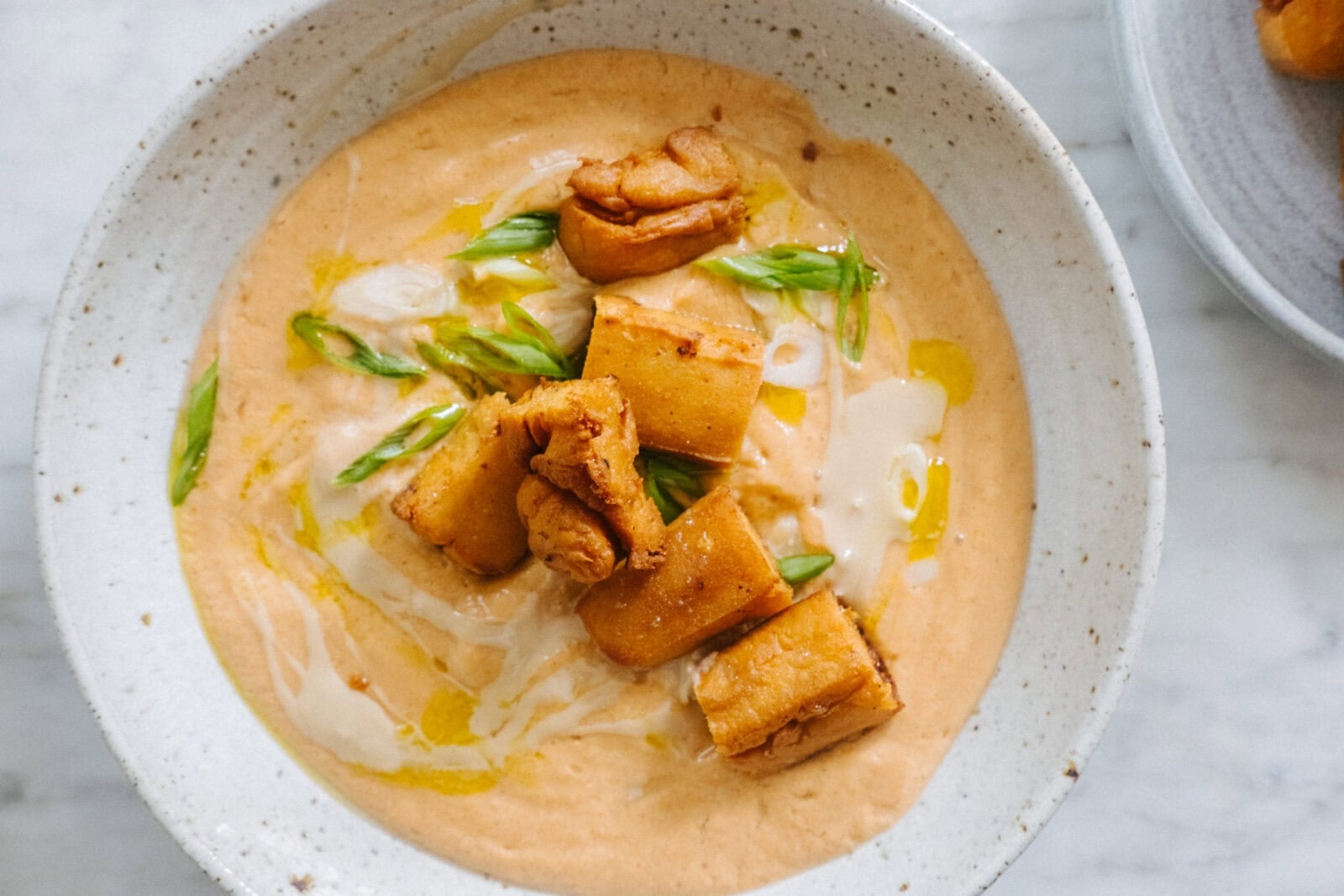 A bowl of creamy orange soup topped with golden fried tofu pieces, green onion slices, a drizzle of oil, and a swirl of cream. - Hetty Lui McKinnon