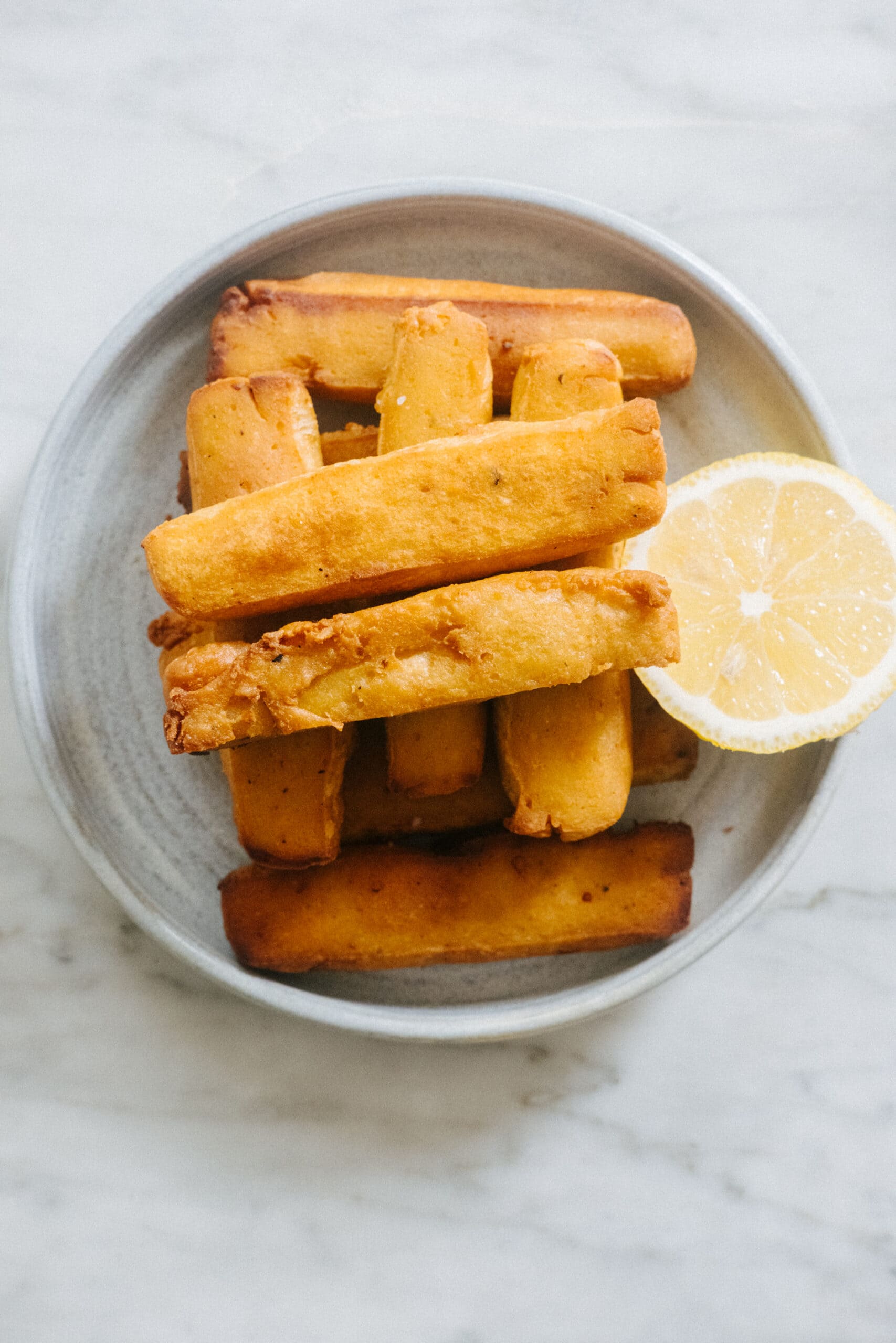 A plate with several golden-brown yuca fries stacked on top of each other and a lemon slice on the side, set on a light marble surface.