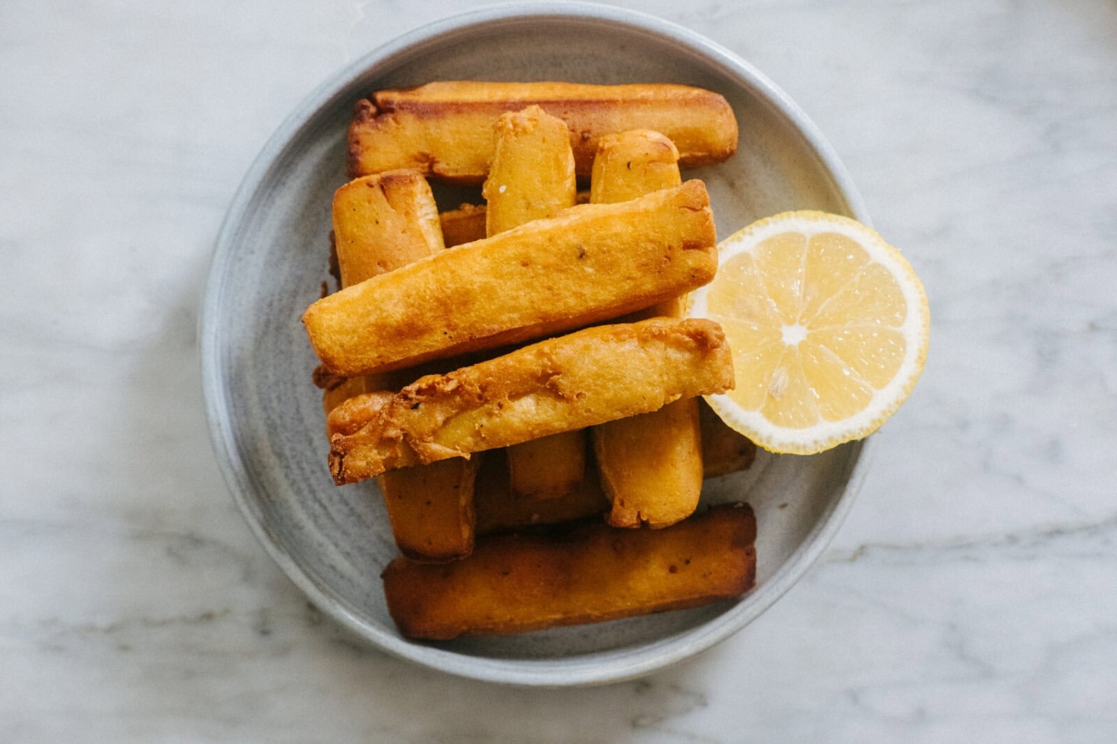 A plate with several golden fried yuca sticks and a sliced lemon on the side, placed on a light marble surface. - Hetty Lui McKinnon