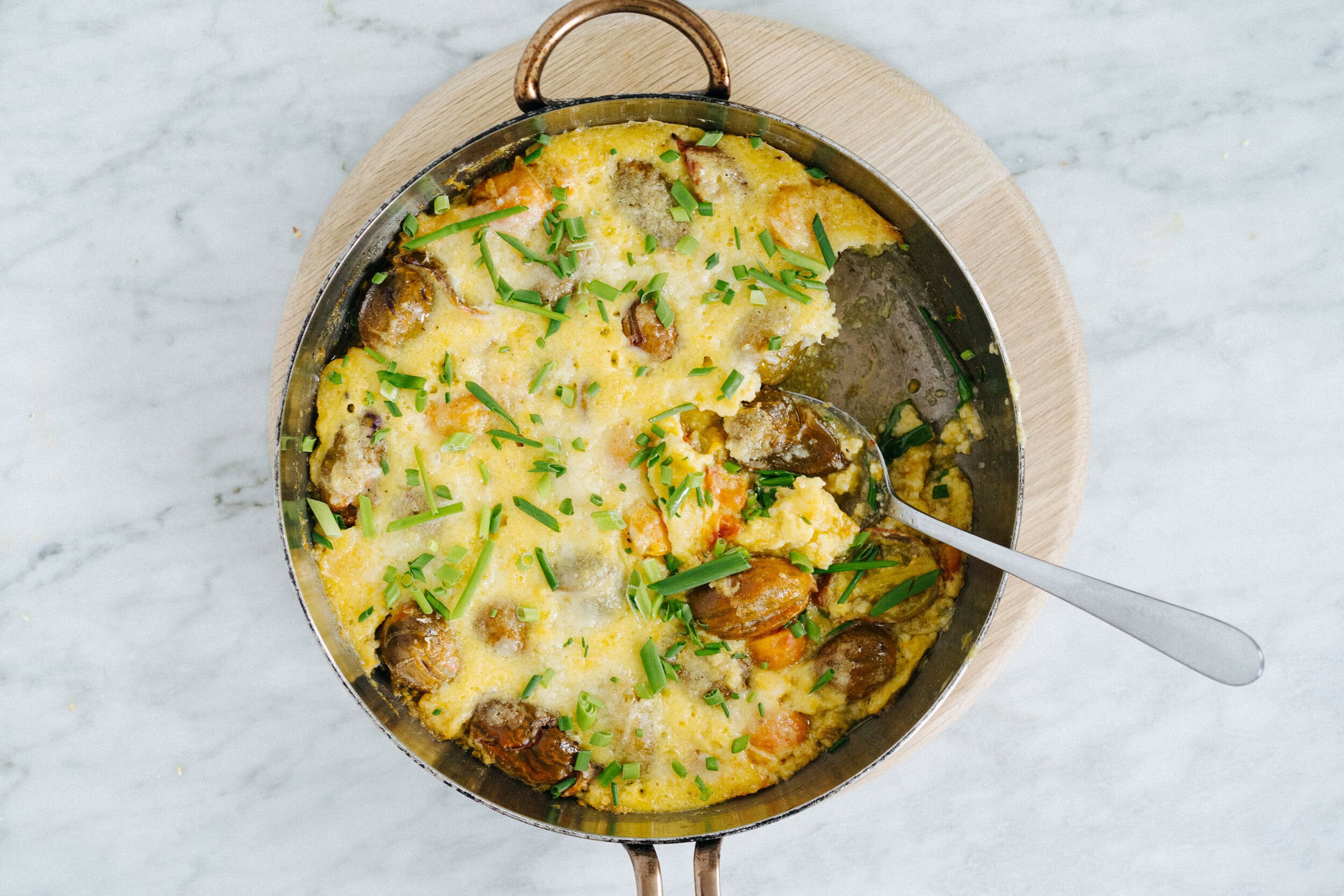 A round pan of baked egg casserole with potatoes, cheese, and chopped herbs, partially served with a spoon resting inside.