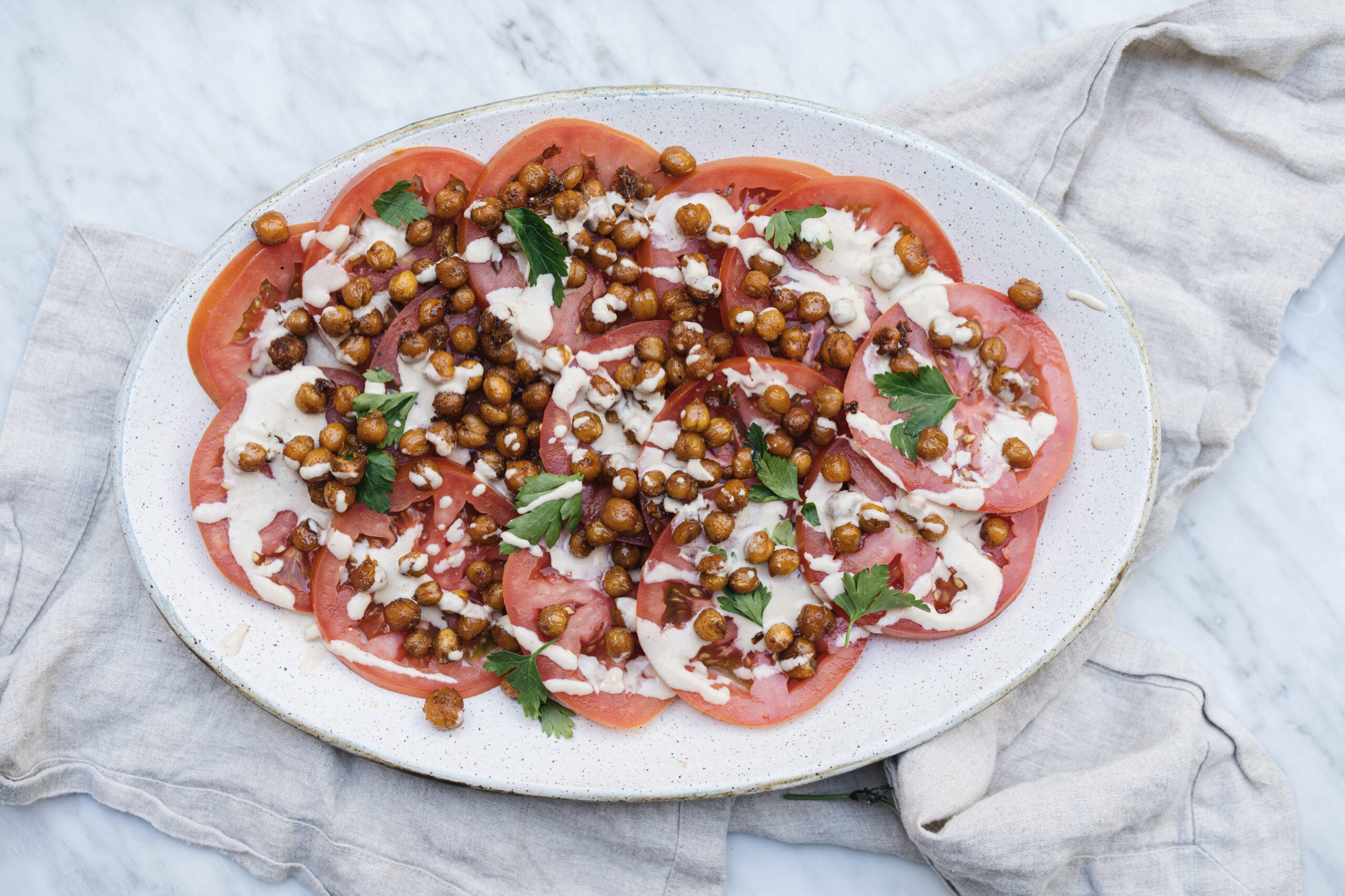 Oval plate with sliced tomatoes, roasted chickpeas, fresh parsley, and a drizzle of white sauce, set on a marble surface with a light-colored cloth underneath. - Hetty Lui McKinnon