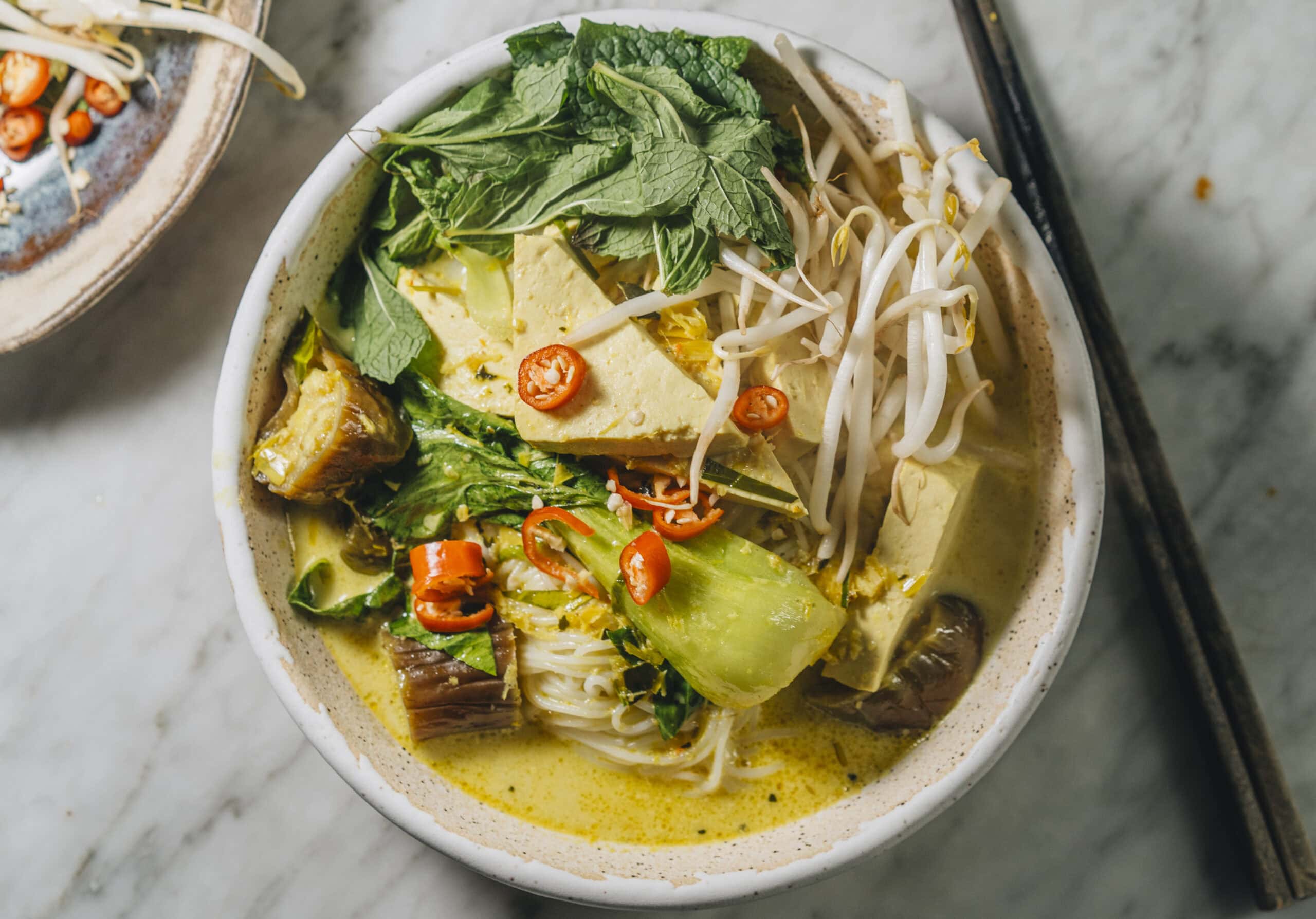 A bowl of yellow curry noodle soup with tofu, eggplant, bok choy, bean sprouts, fresh herbs, and sliced red chili on top.