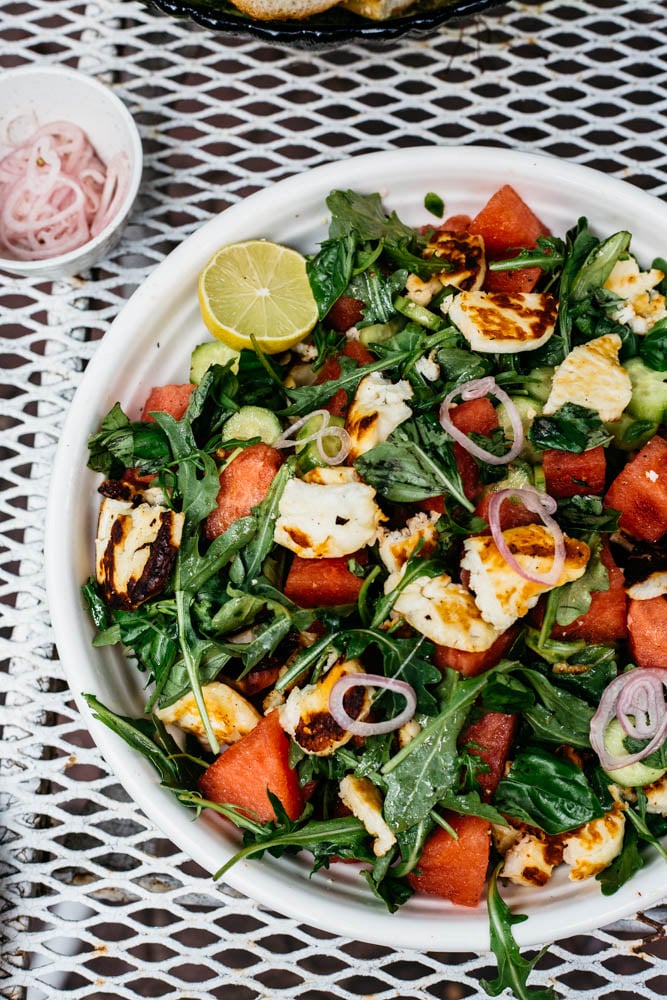 A bowl of salad with watermelon, grilled halloumi cheese, arugula, cucumber, and sliced red onions, garnished with a lime wedge, sits on a white metal table. - Hetty Lui McKinnon