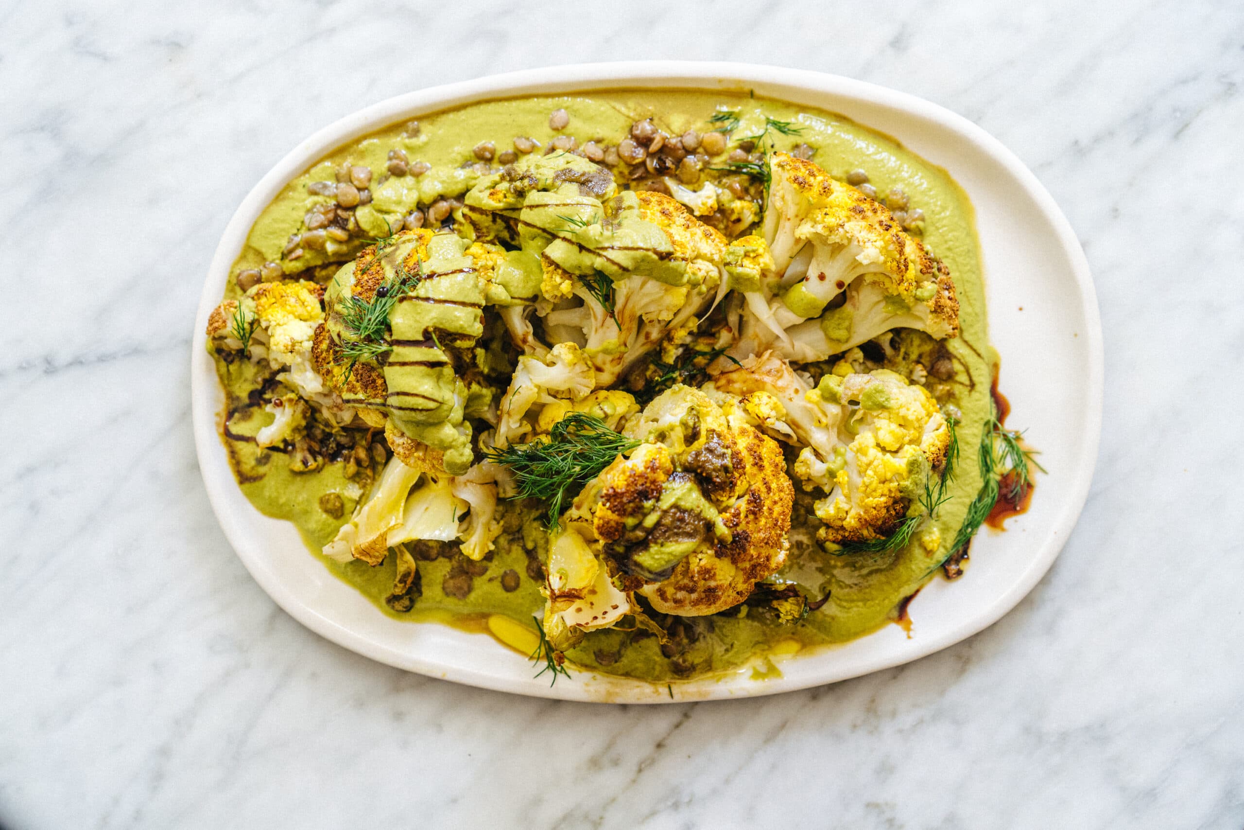 Roasted cauliflower served on a white oval plate with green sauce, lentils, fresh herbs, and a drizzle of dark balsamic glaze.