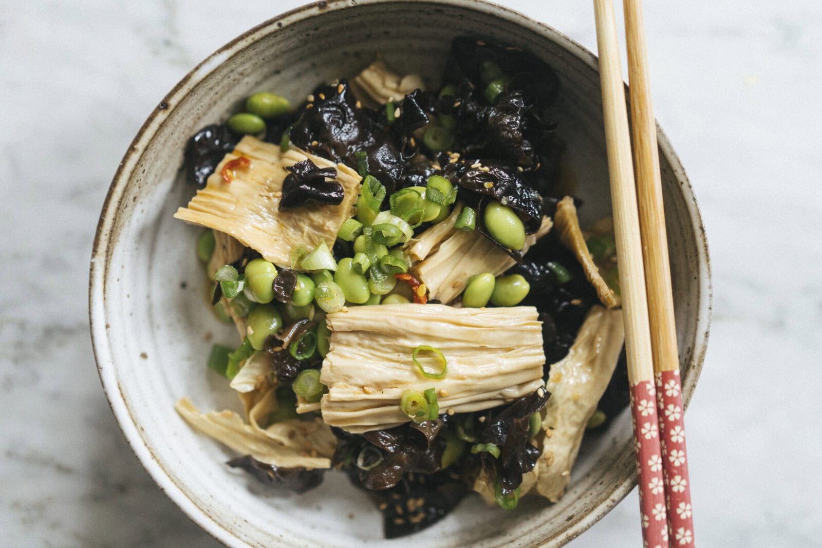 A bowl of tofu skin salad with black fungus, edamame, chopped green onions, and chopsticks resting on the rim.
