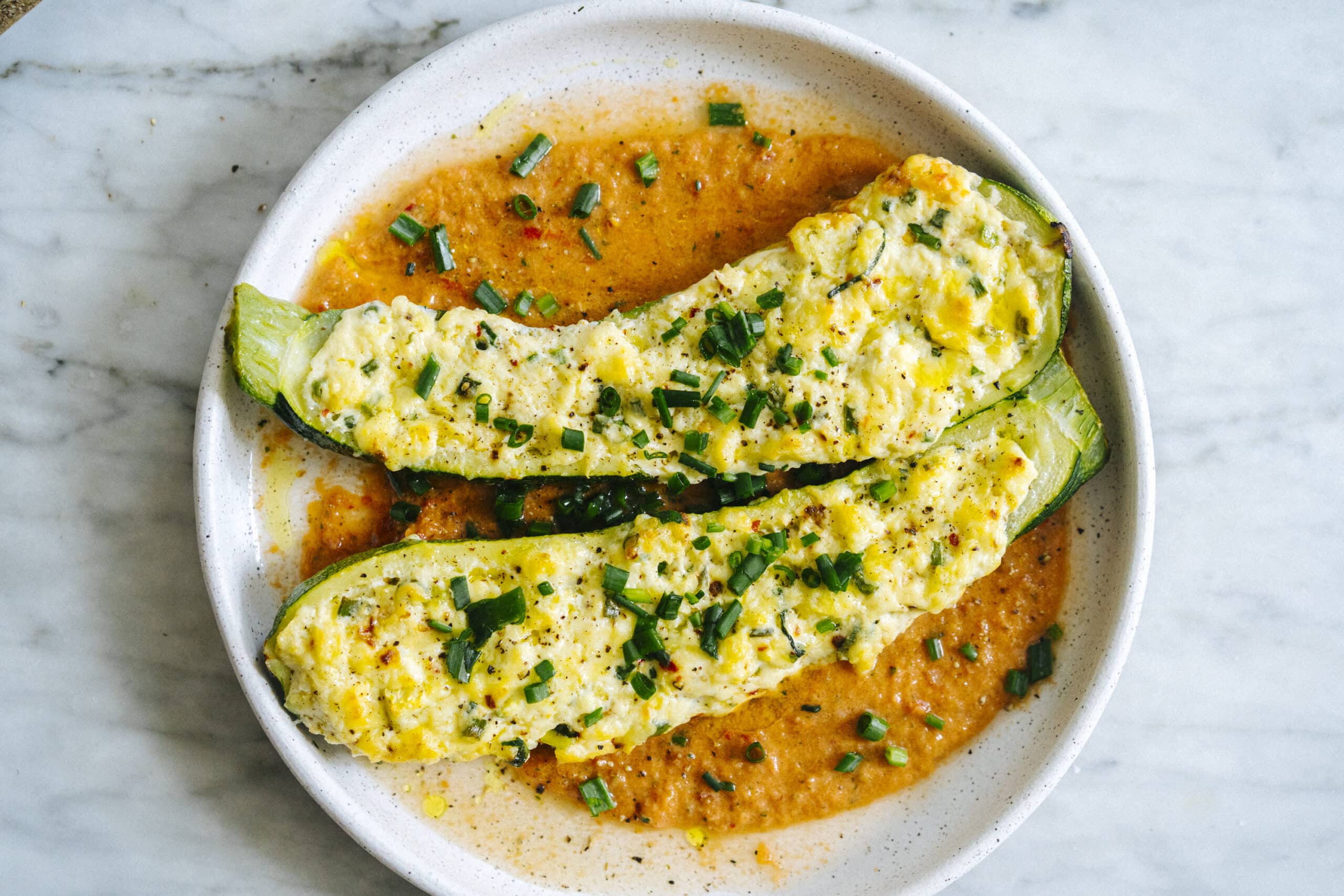 Two zucchini halves stuffed with a cheesy filling are served on a white plate over a tomato-based sauce, garnished with chopped green onions and black pepper.