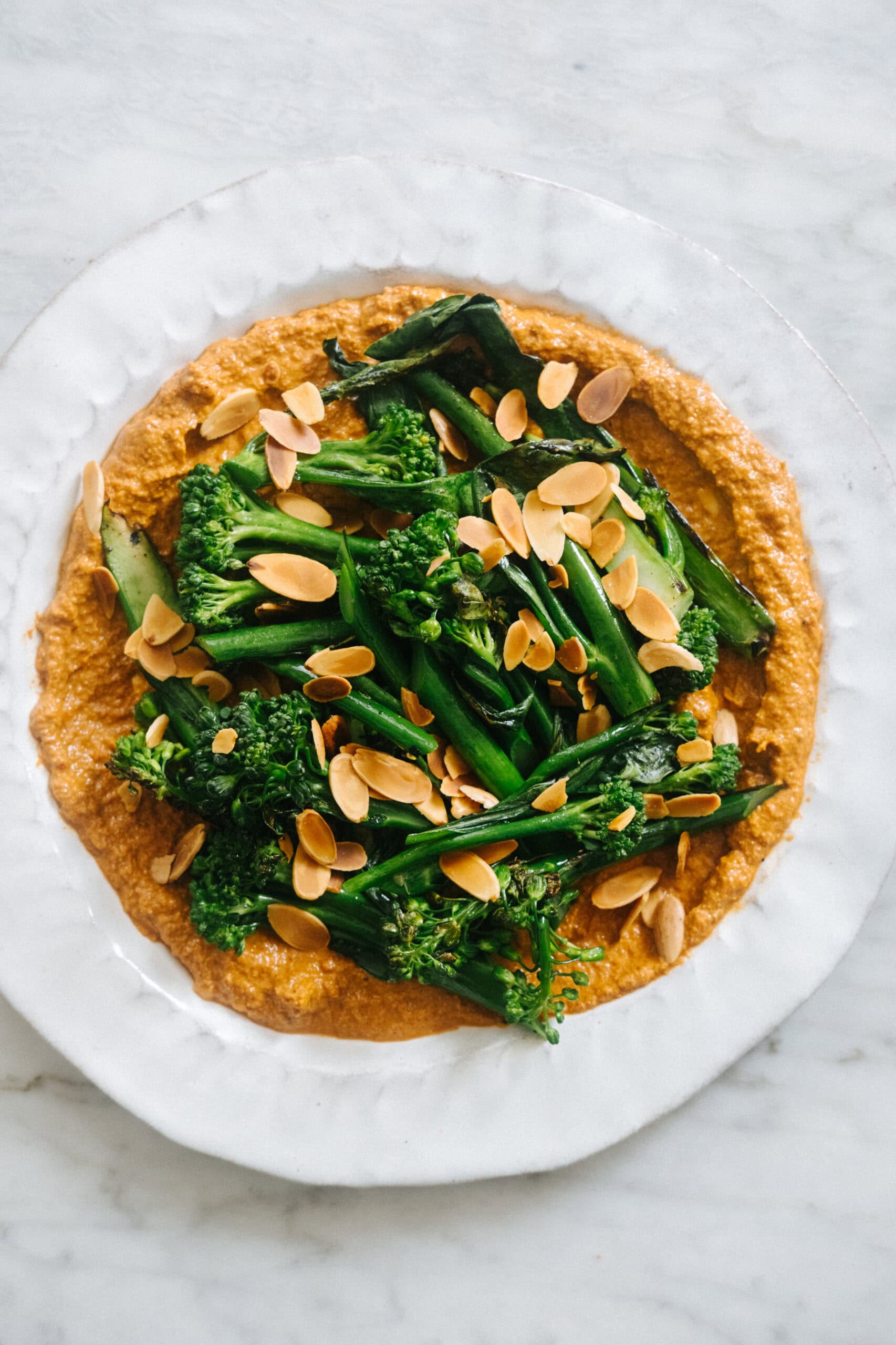A white plate with broccolini and toasted almond slices on a spread of reddish-brown dip, placed on a marble surface.