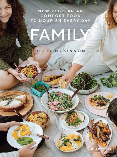 Four people sit on a picnic blanket sharing various vegetarian dishes, including salads, roasted vegetables, and fruit, with the book title "Family" by Hetty McKinnon displayed.