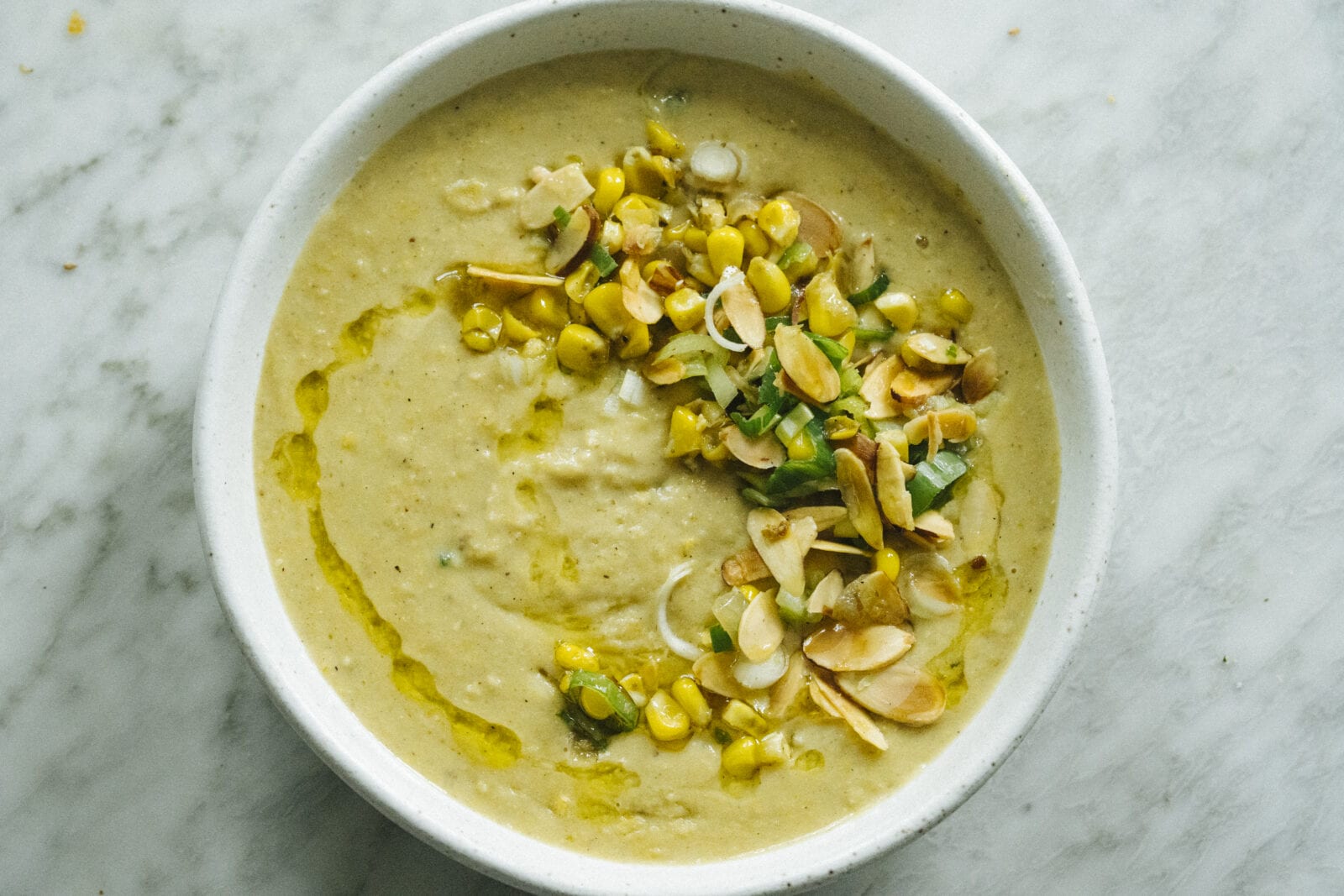 A bowl of creamy soup topped with corn, sliced almonds, chopped green herbs, and a drizzle of oil, on a marble surface. - Hetty Lui McKinnon