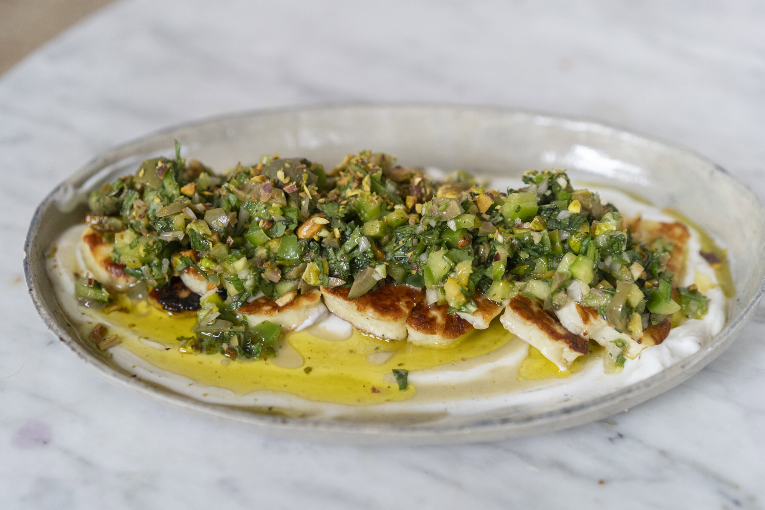 Grilled cheese slices topped with a green herb and vegetable salsa on a bed of creamy sauce, drizzled with olive oil, served on an oval plate.