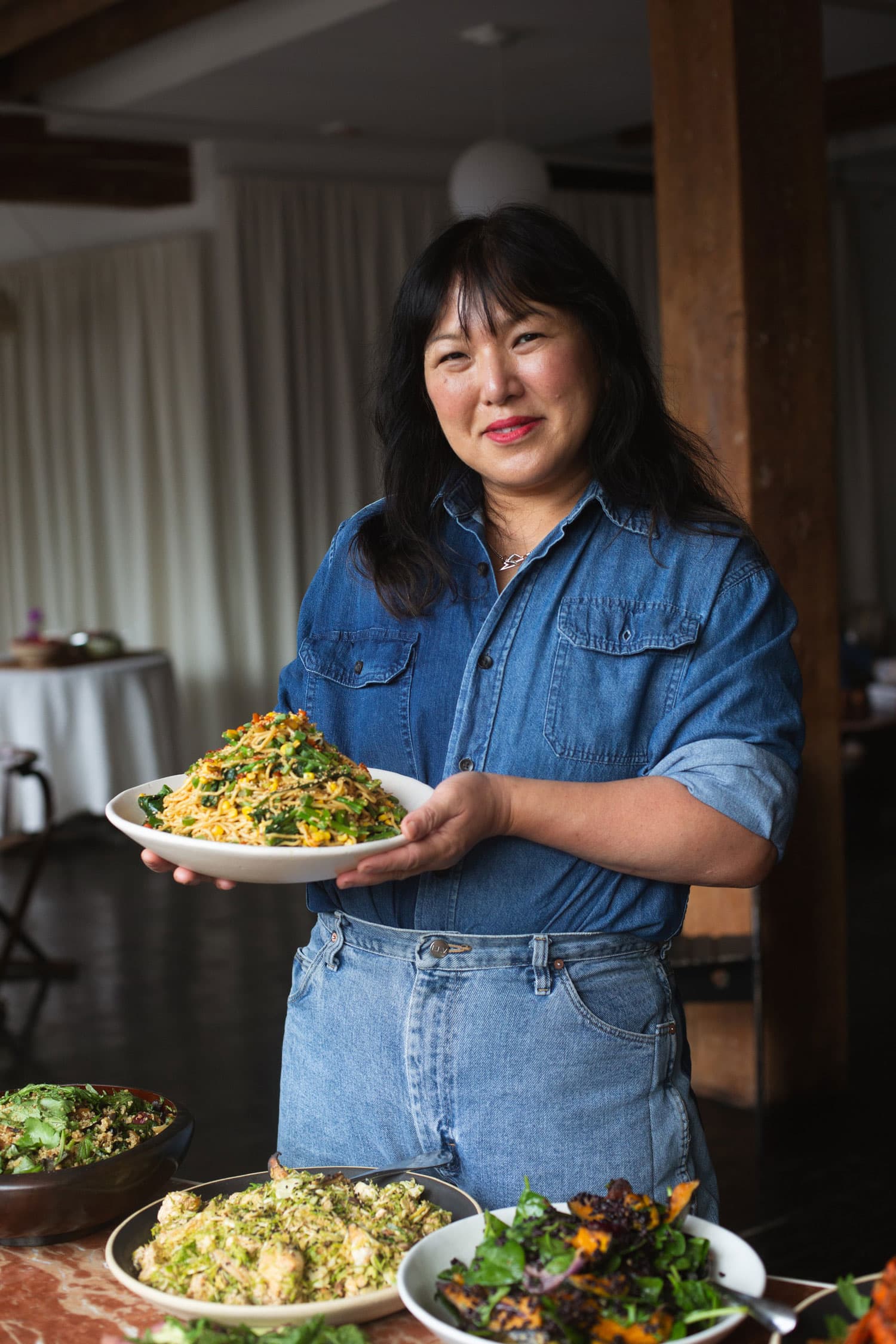 A woman in a denim shirt and jeans holds a plate of food, standing behind a table with several dishes in a warmly lit room.