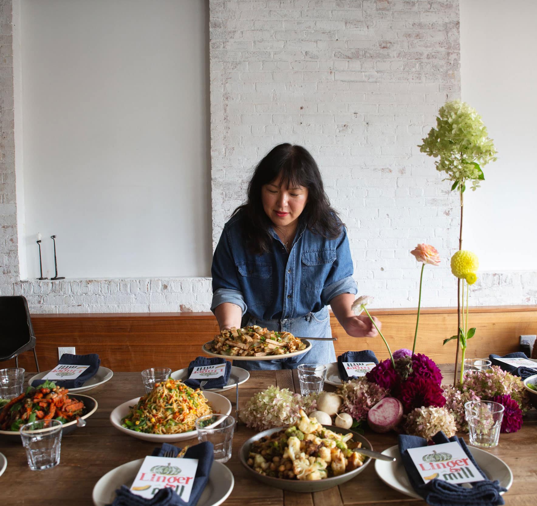 A woman in a denim shirt sets a dish on a table filled with various plates of food, flowers, and "Linger longer" cards in a bright, modern dining space.