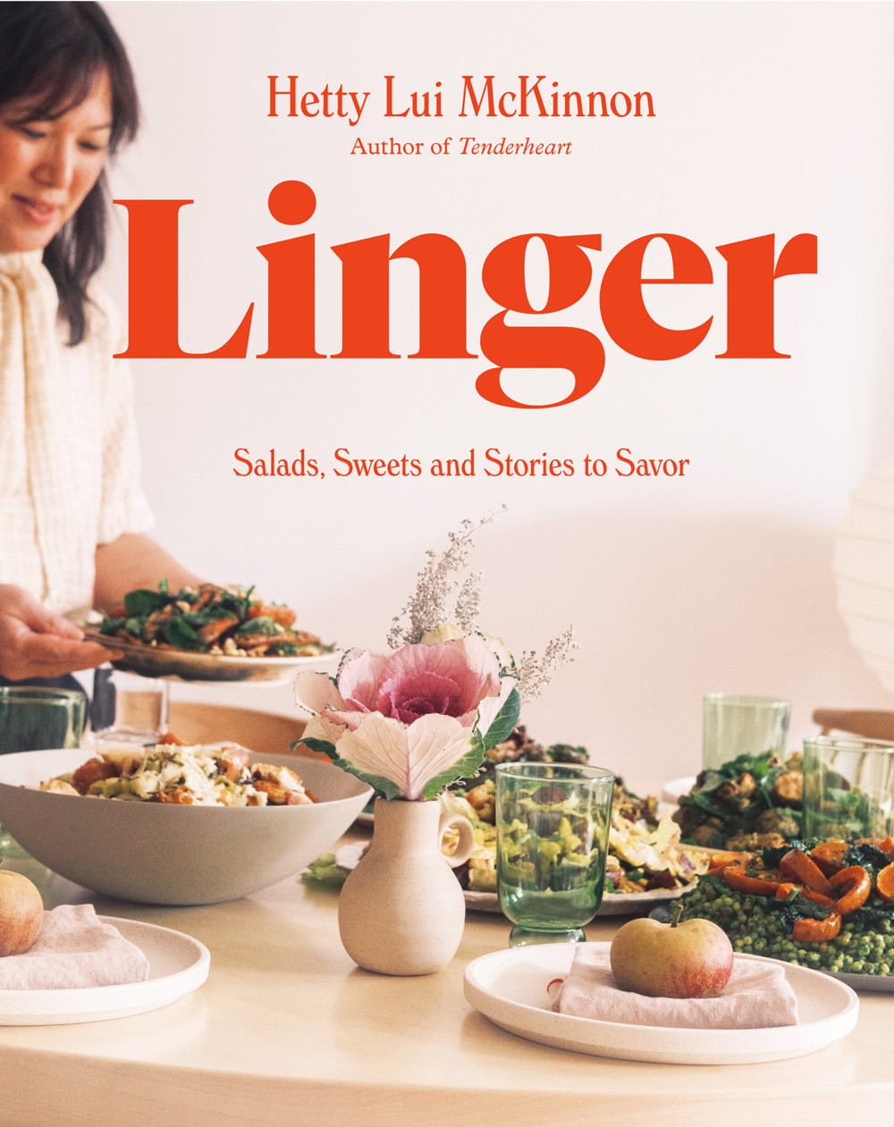 A woman sets a dining table with plates of food, flowers, fruit, and drinks. The book cover reads "Linger: Salads, Sweets and Stories to Savor" by Hetty Lui McKinnon.