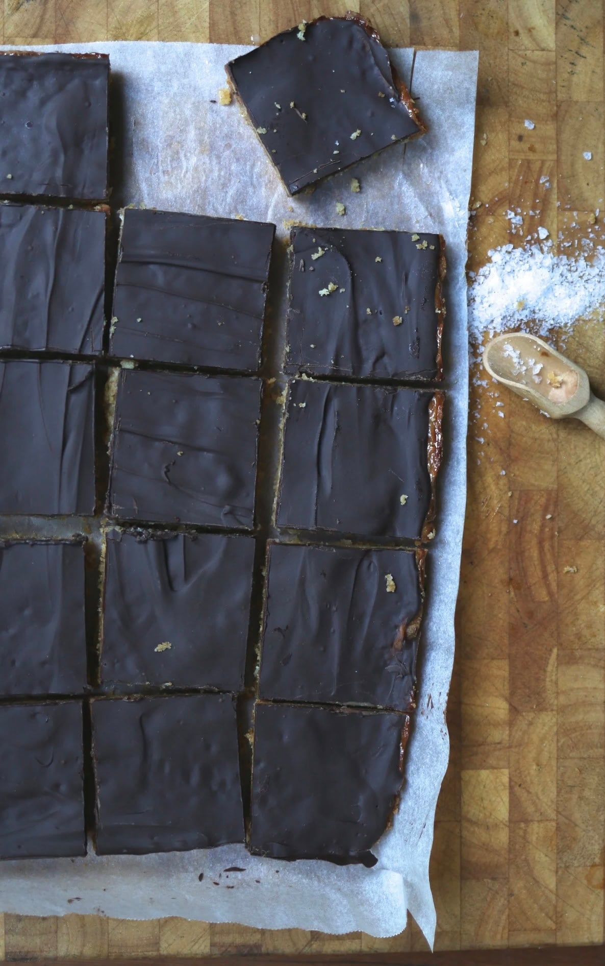 A tray of chocolate-topped bars cut into squares on parchment paper, with a wooden spoon and scattered powdered sugar on a wooden surface. - Hetty Lui McKinnon