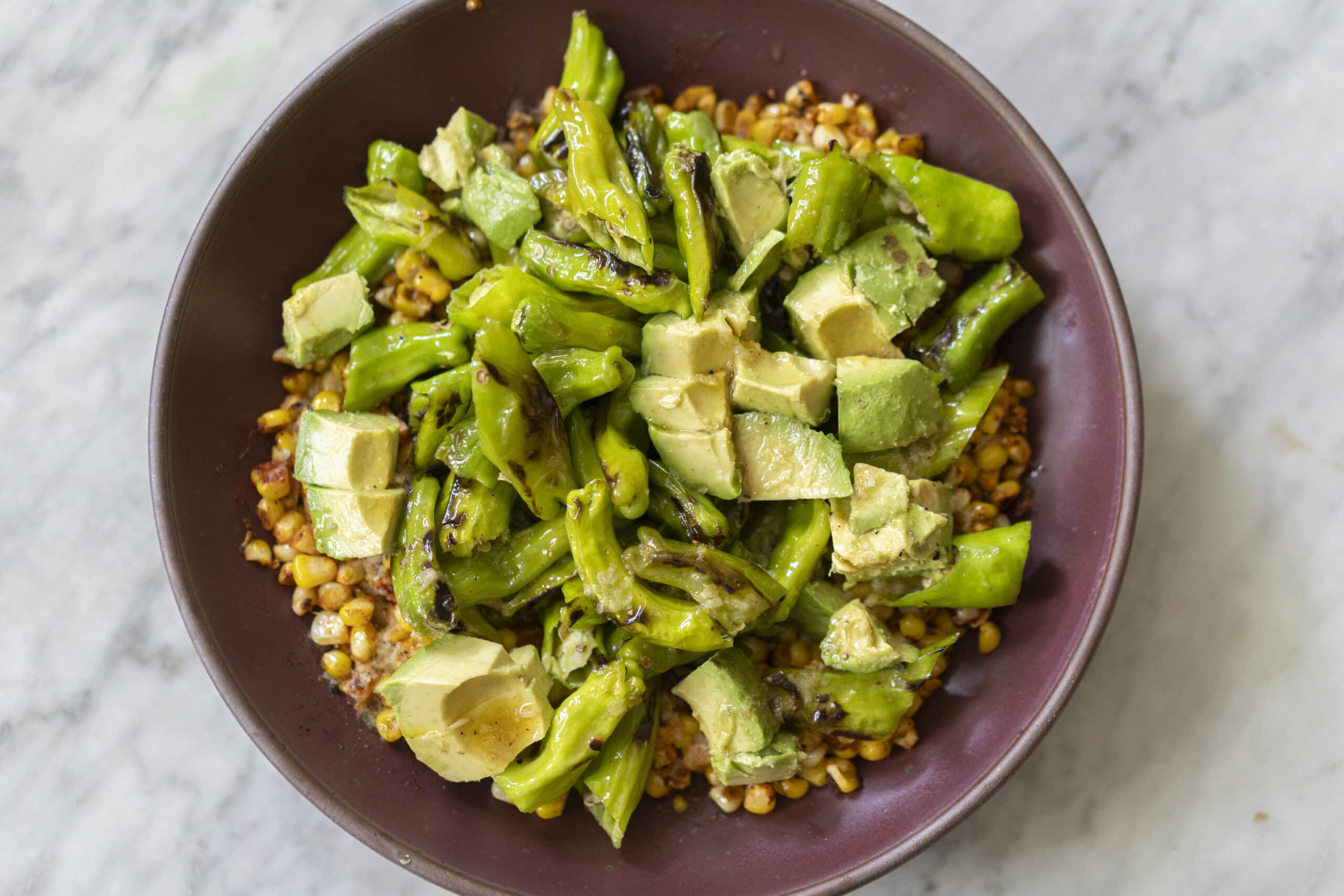 A bowl of roasted corn topped with sliced avocado and cooked green peppers, served on a marble surface.