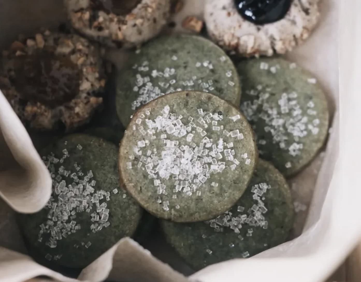 A close-up of round green cookies sprinkled with coarse sugar, surrounded by assorted thumbprint cookies in a parchment-lined container.