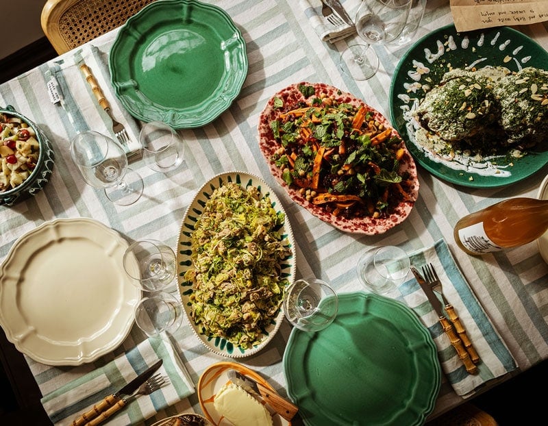 A table set for a meal with green and cream plates, salad dishes, glassware, wine, and a striped tablecloth.