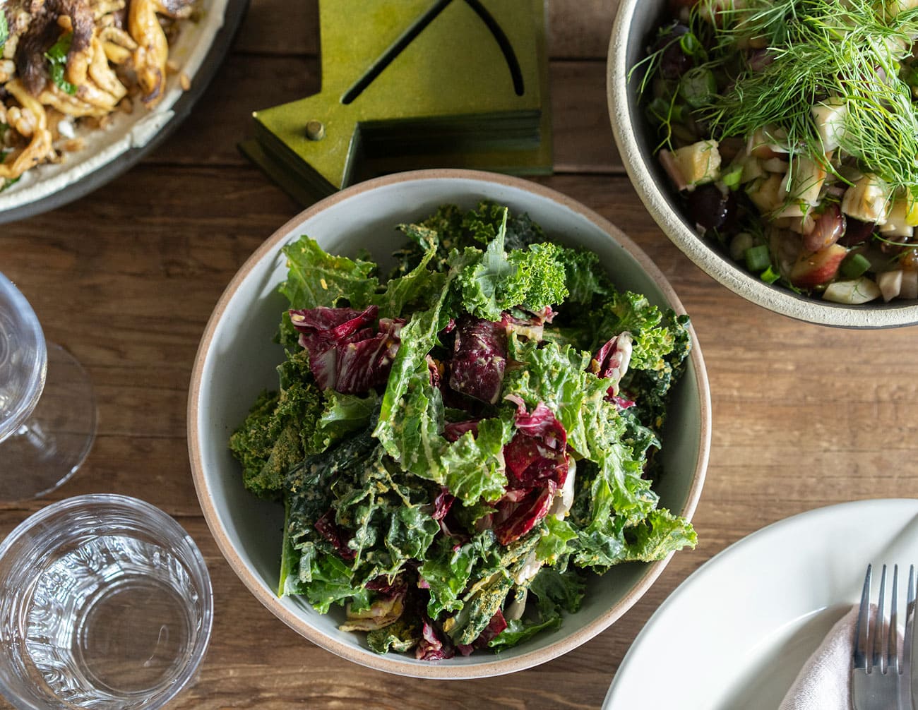 A bowl of mixed leafy green salad sits on a wooden table next to a glass of water, a bowl of chopped vegetables, and an empty plate with a fork and knife.